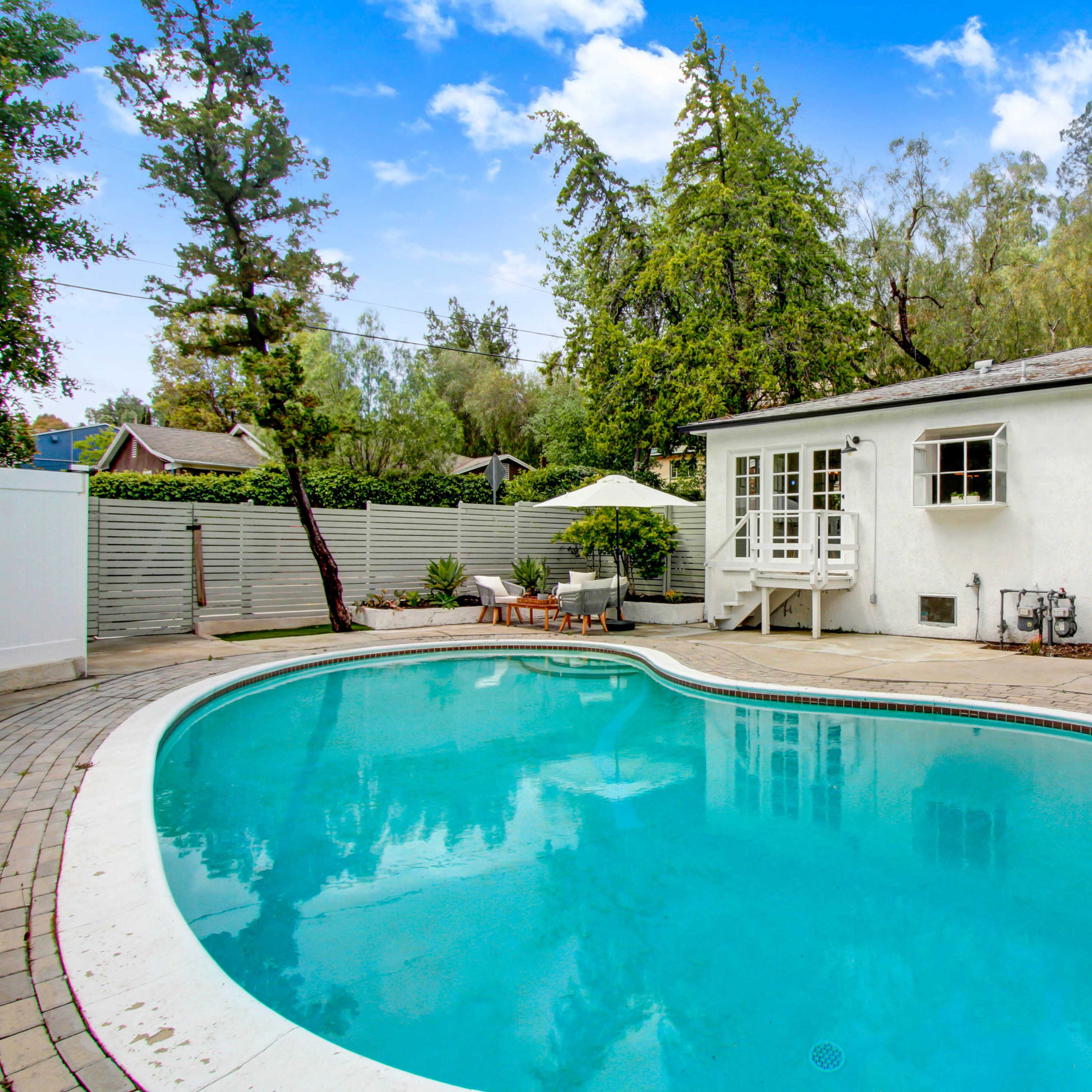 A rectangular swimming pool in a backyard setting, surrounded by trees and a light-colored house with a small deck.