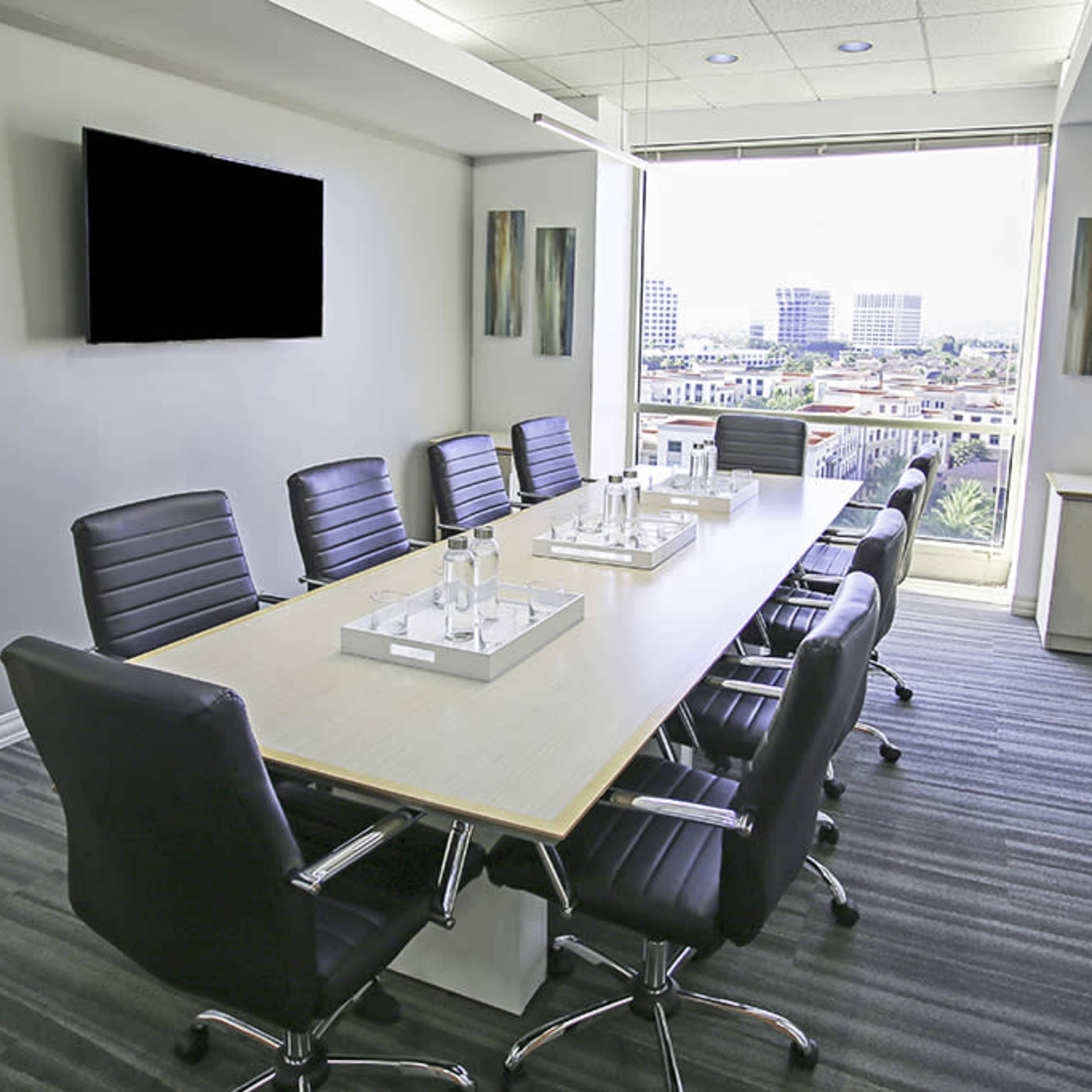 A modern conference room features a long table surrounded by black leather chairs, with a large window offering a view of the city.