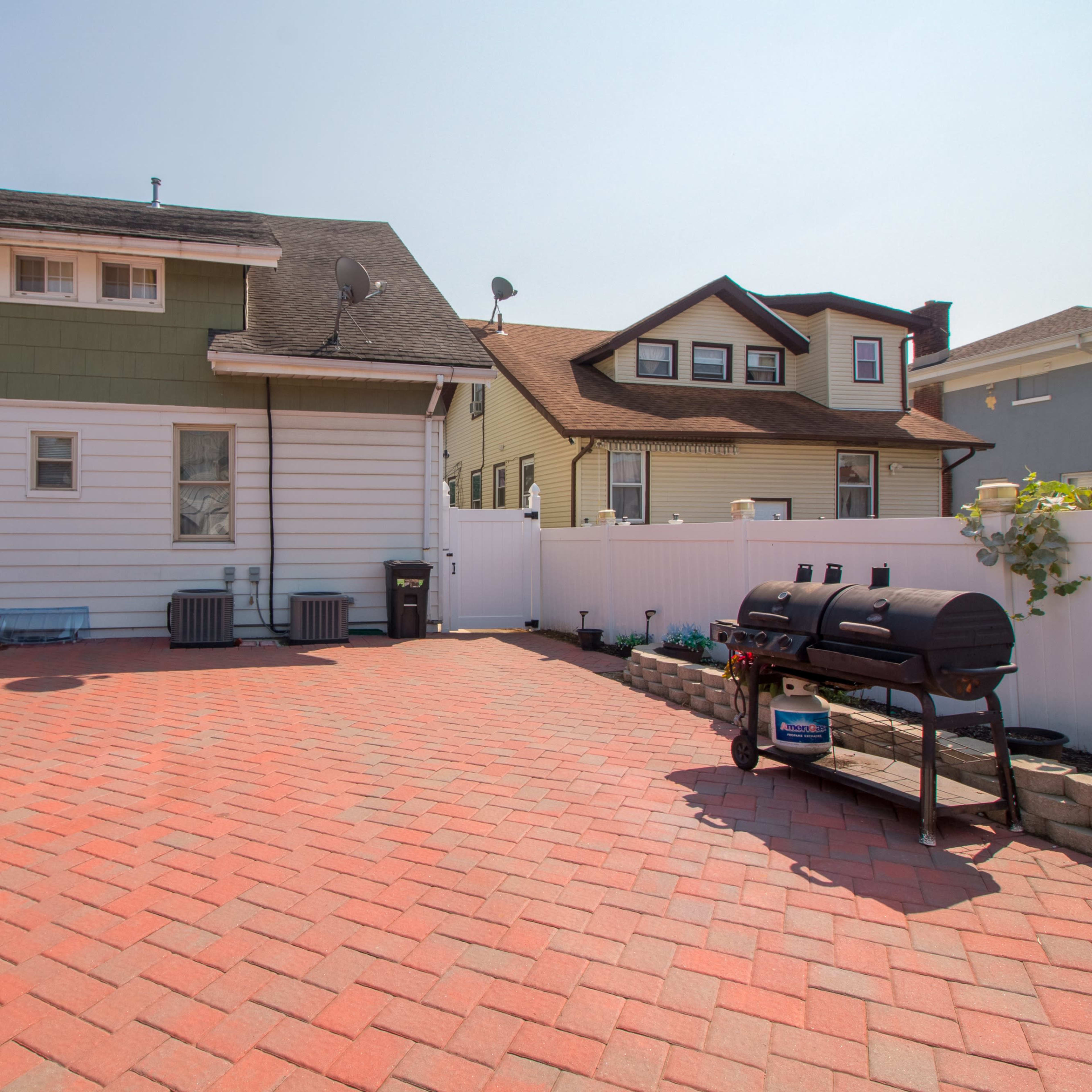 A brick-paved backyard with a barbecue grill, a smoker, and two houses in the background.