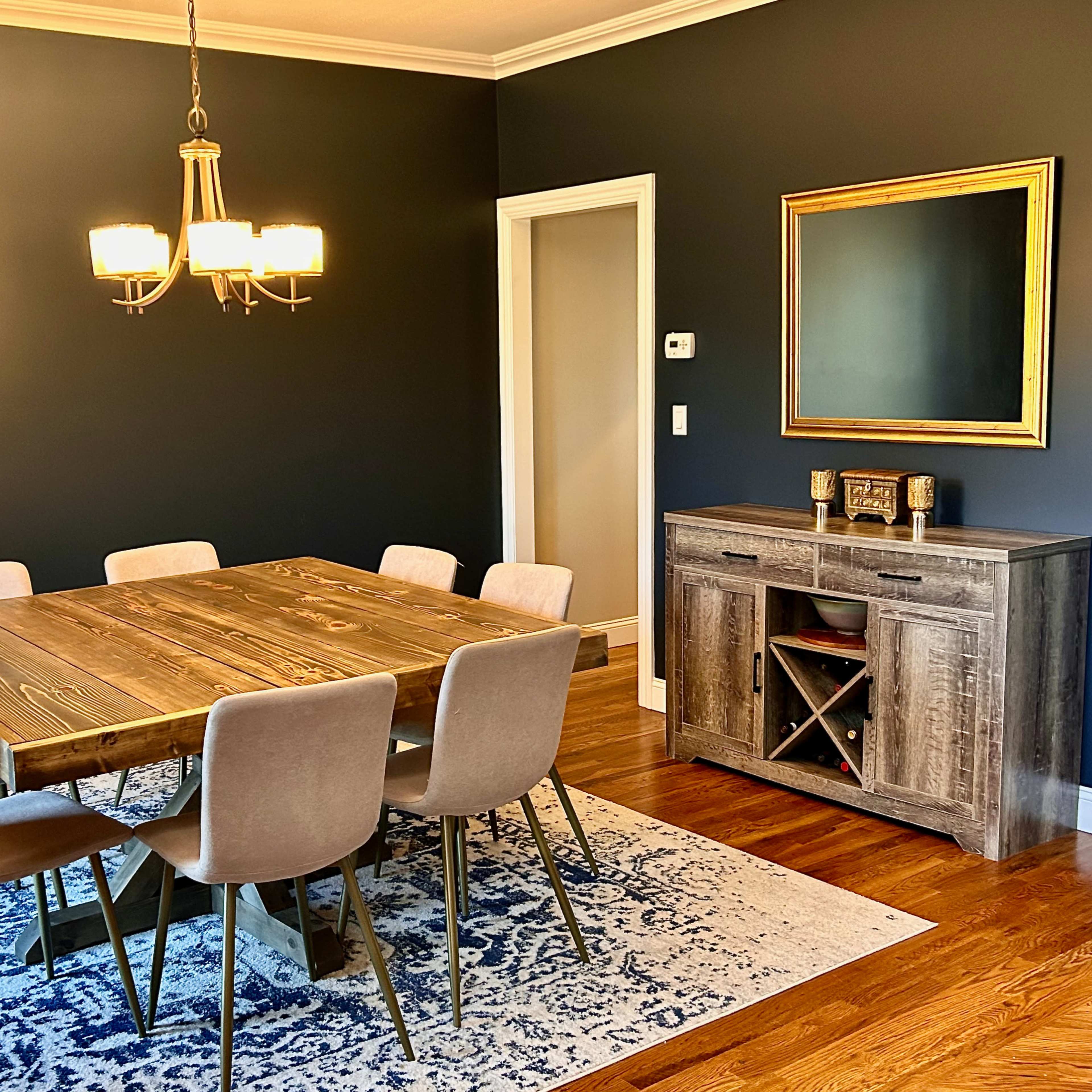 A dining room featuring a wooden table with eight chairs, a sideboard with wine storage, and a decorative mirror on a dark wall.