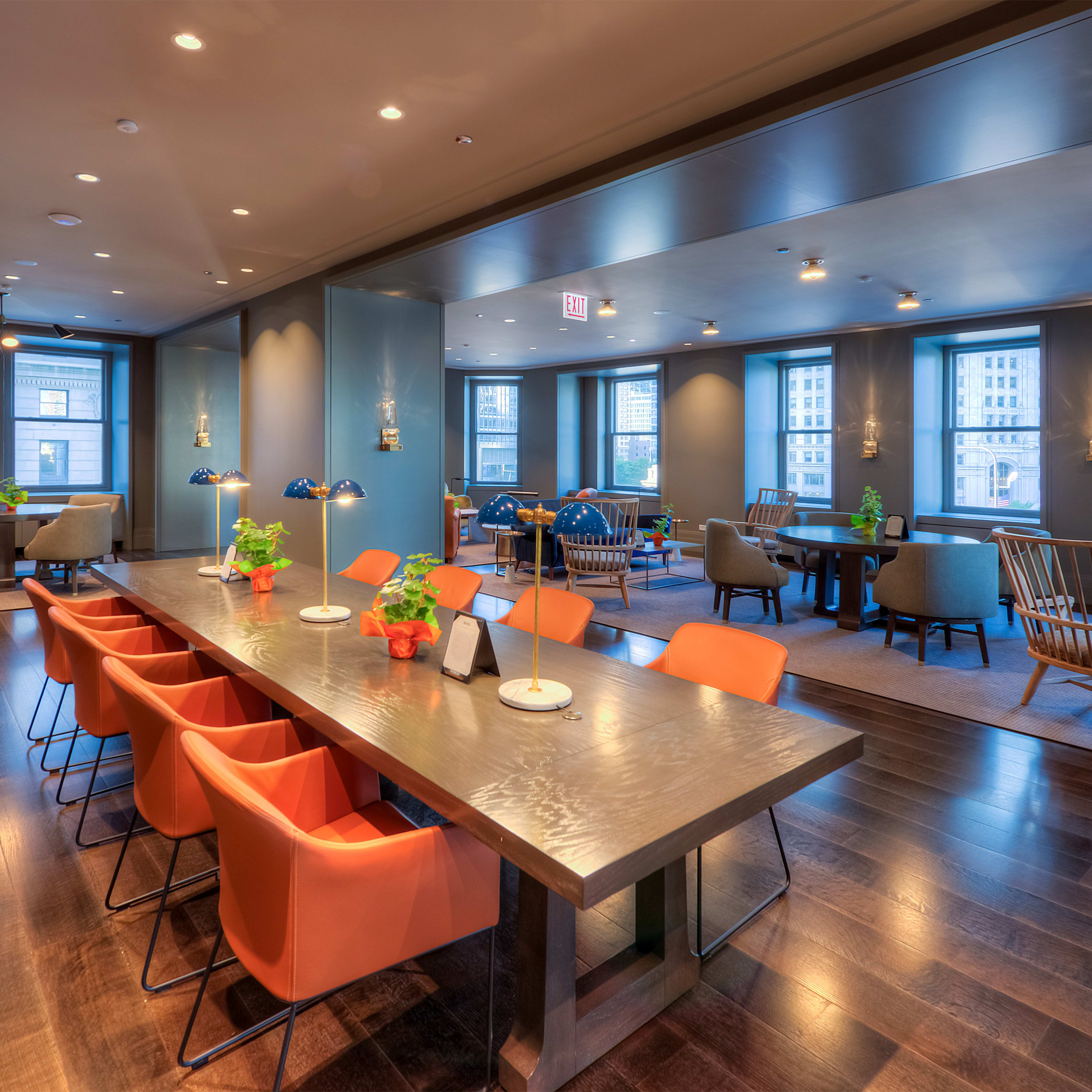 A modern room with a long dining table surrounded by orange chairs, alongside seating areas with plants and large windows letting in natural light.