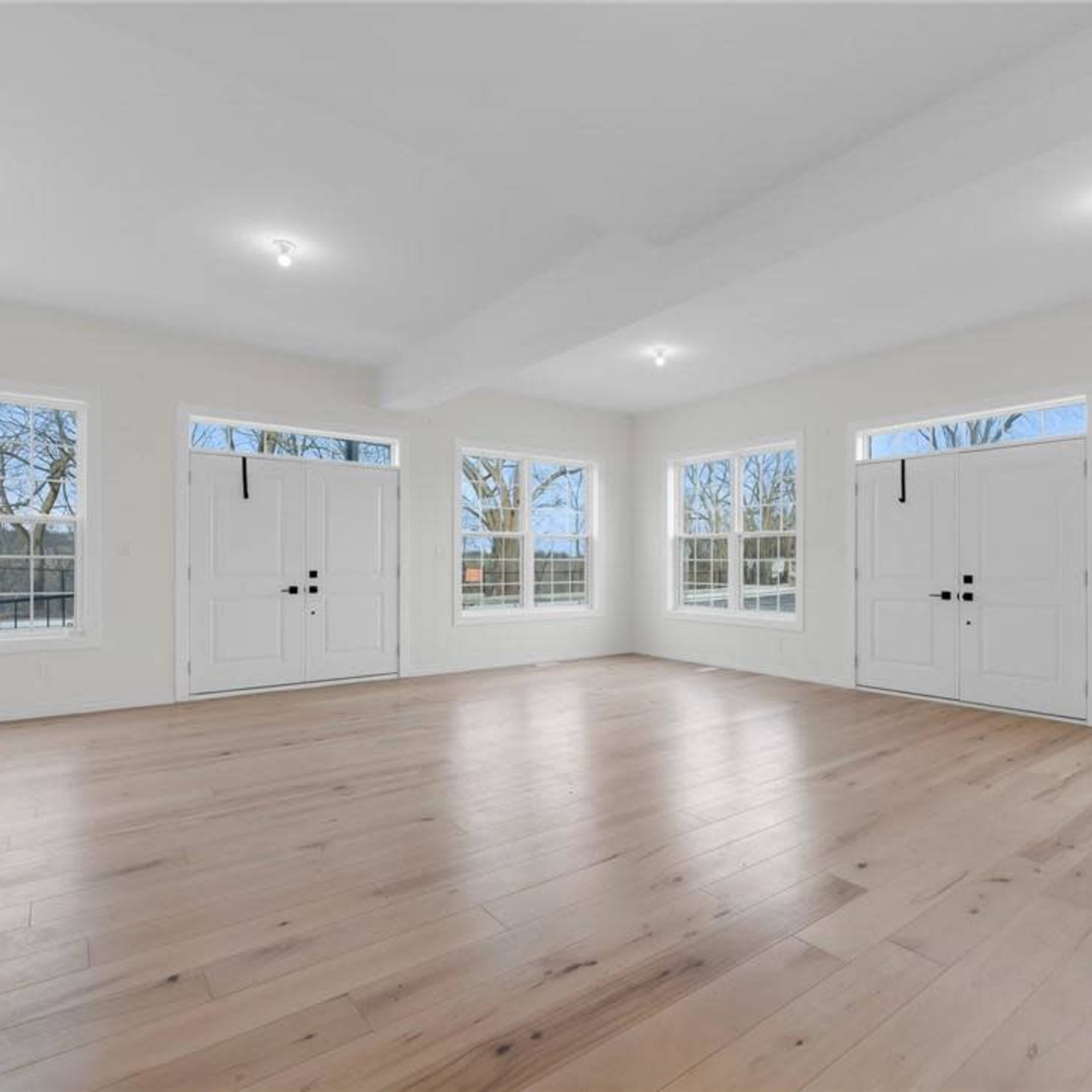 An empty room with wooden floors, large windows, and double doors, allowing natural light to enter.
