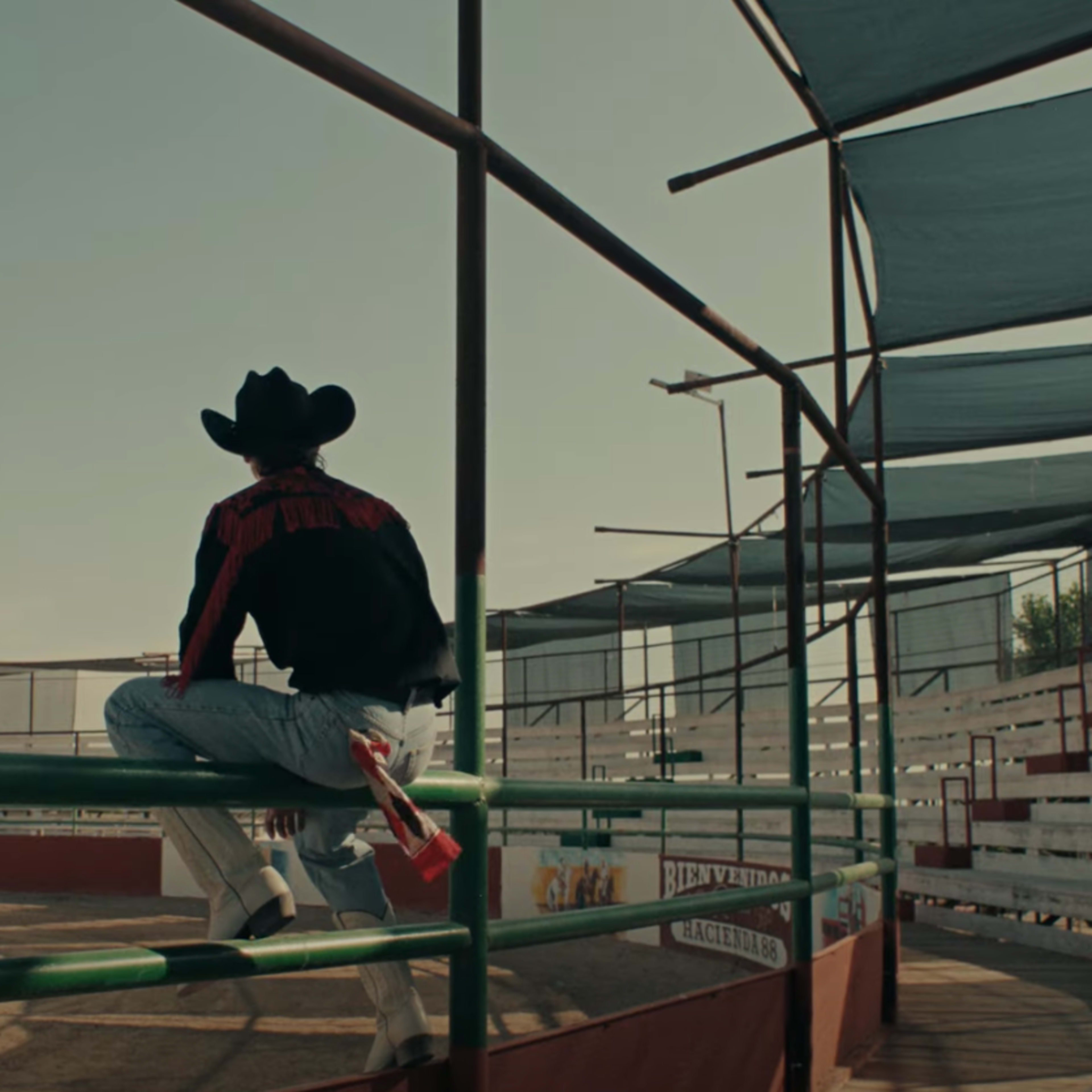 A man in a cowboy hat sits on a railing overlooking an empty arena.