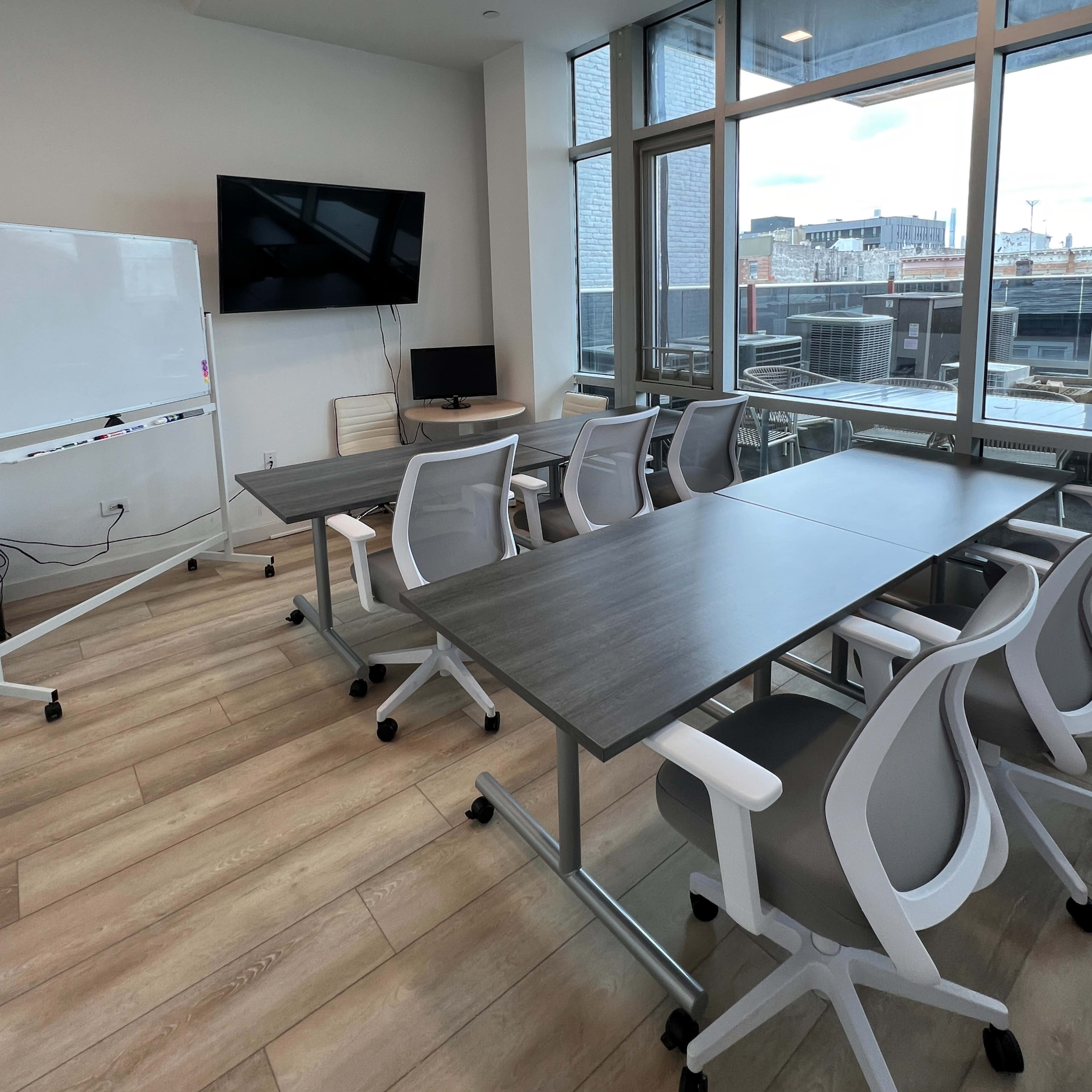 A modern conference room features multiple tables and ergonomic chairs, with a whiteboard and large screen mounted on the wall.