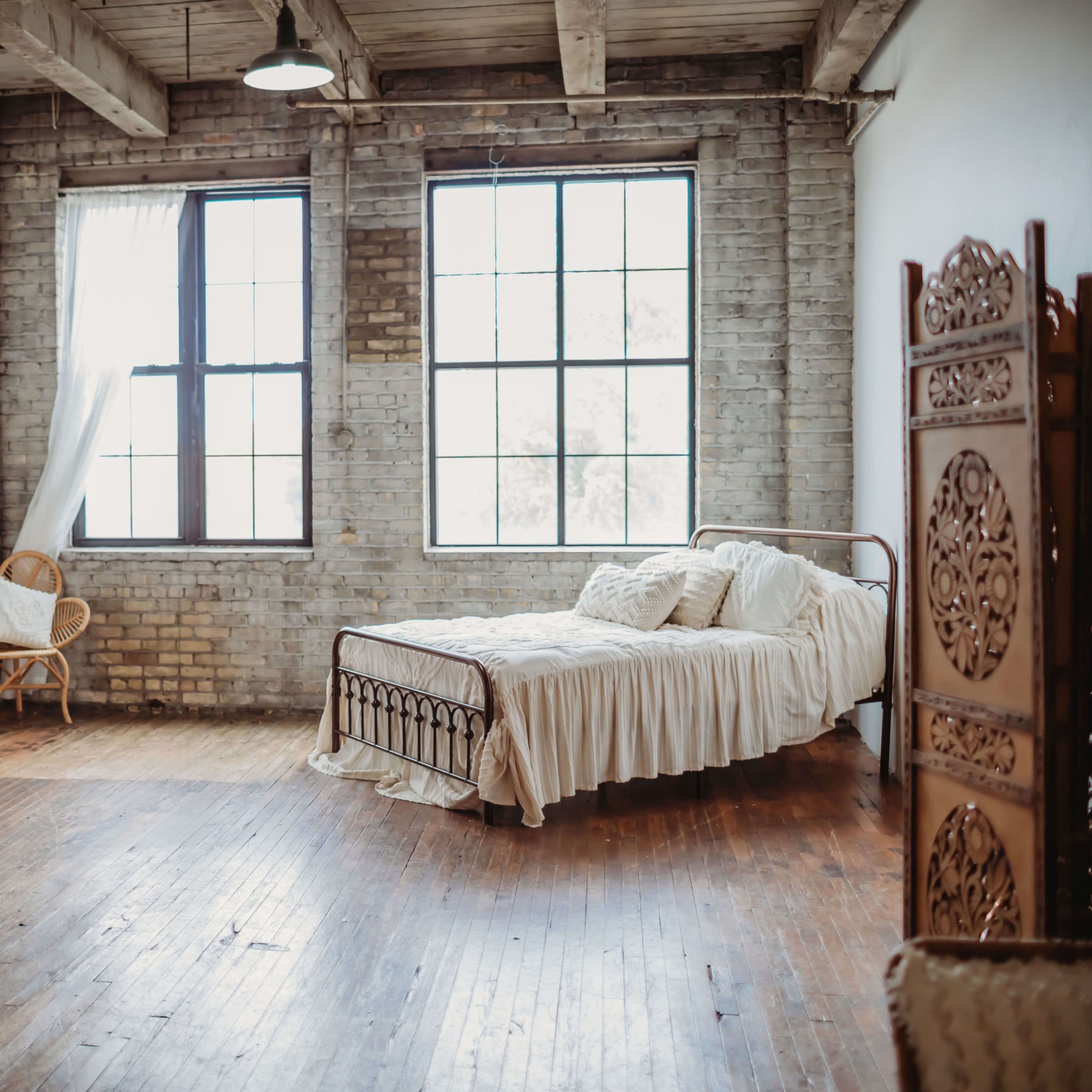 A spacious bedroom with large windows, a metal bed with a white bedspread, and wicker furniture arranged around a wooden floor.