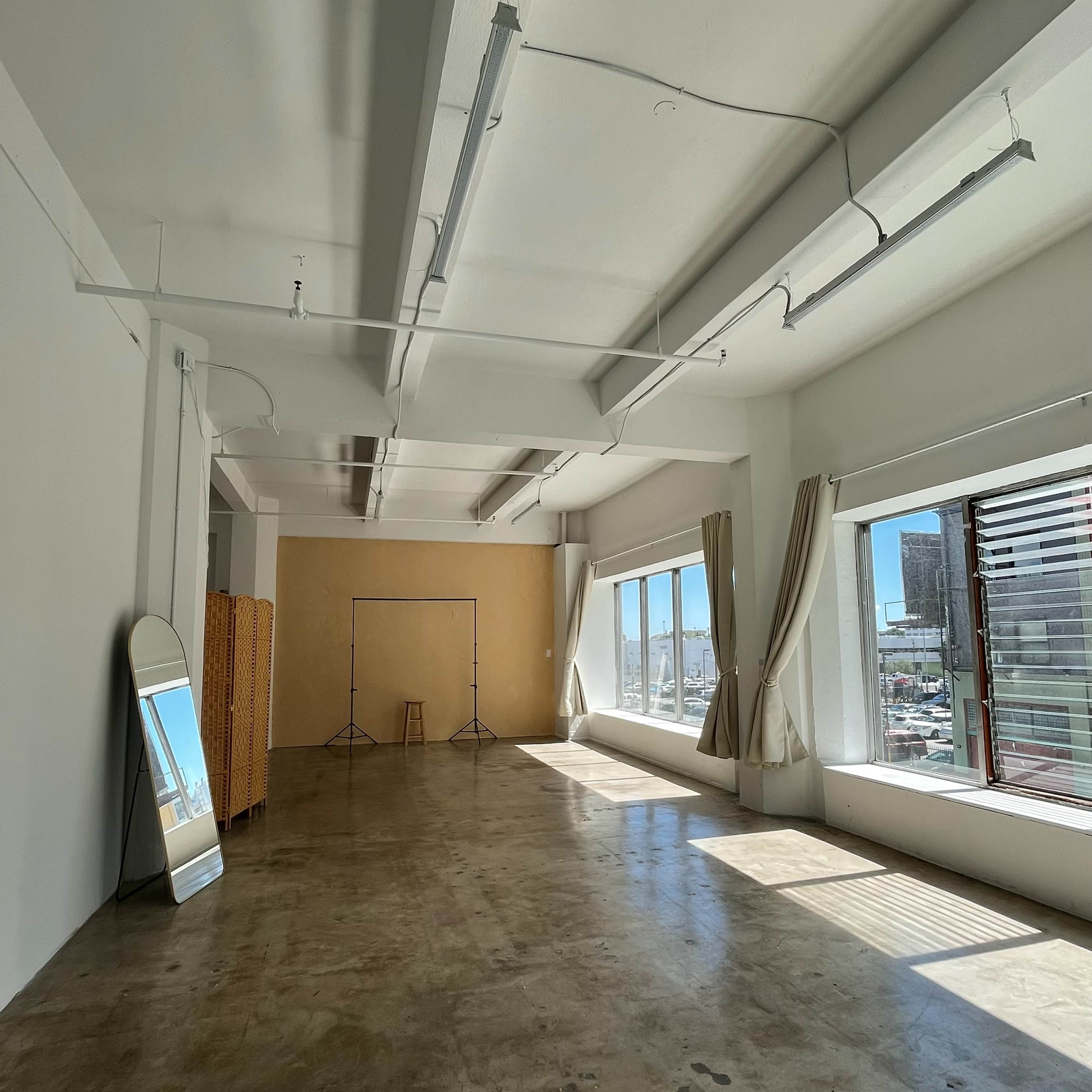 An empty, spacious room with large windows, a mirror, and a plain backdrop setup.