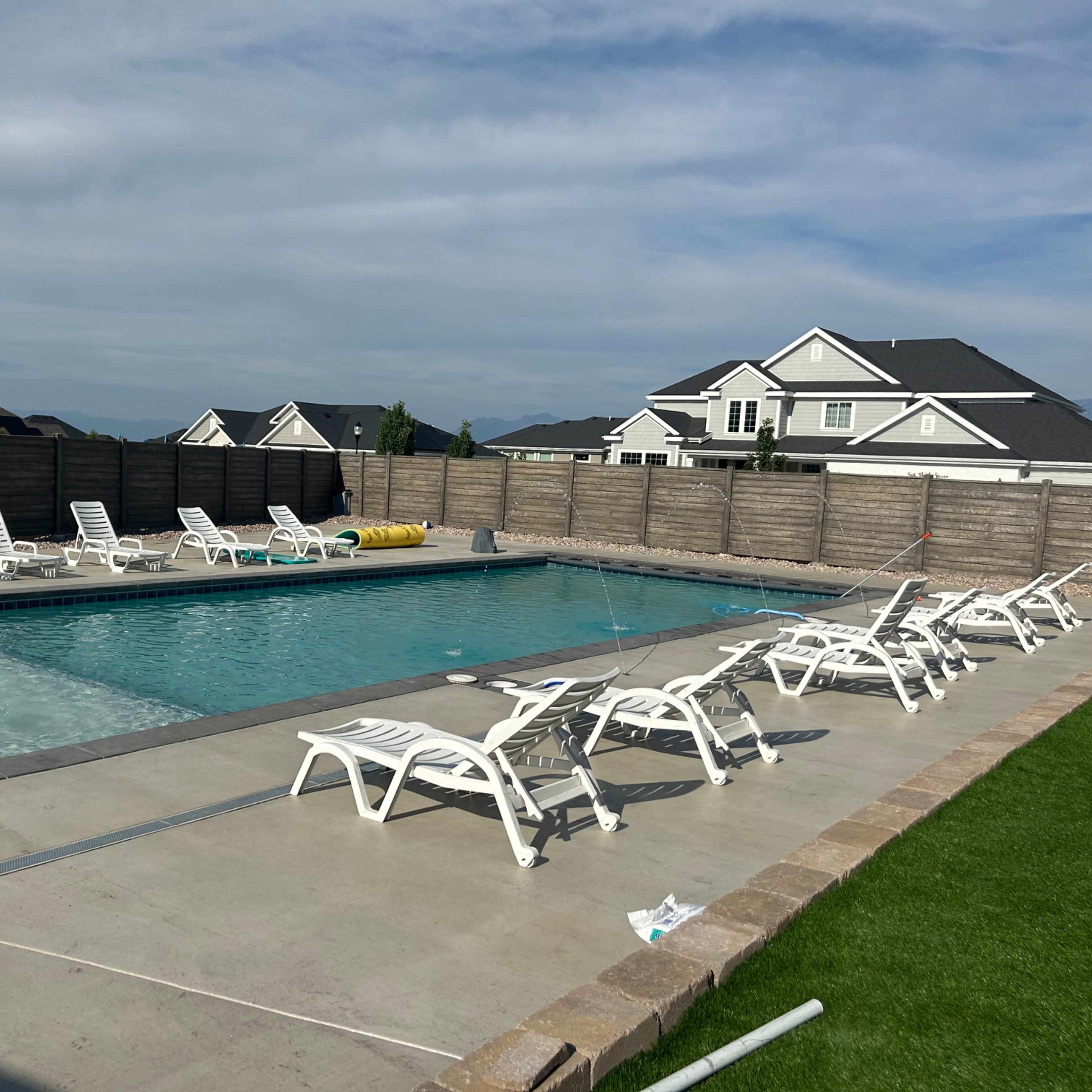 A rectangular swimming pool is surrounded by lounge chairs on a concrete deck, with a wooden fence and houses visible in the background.