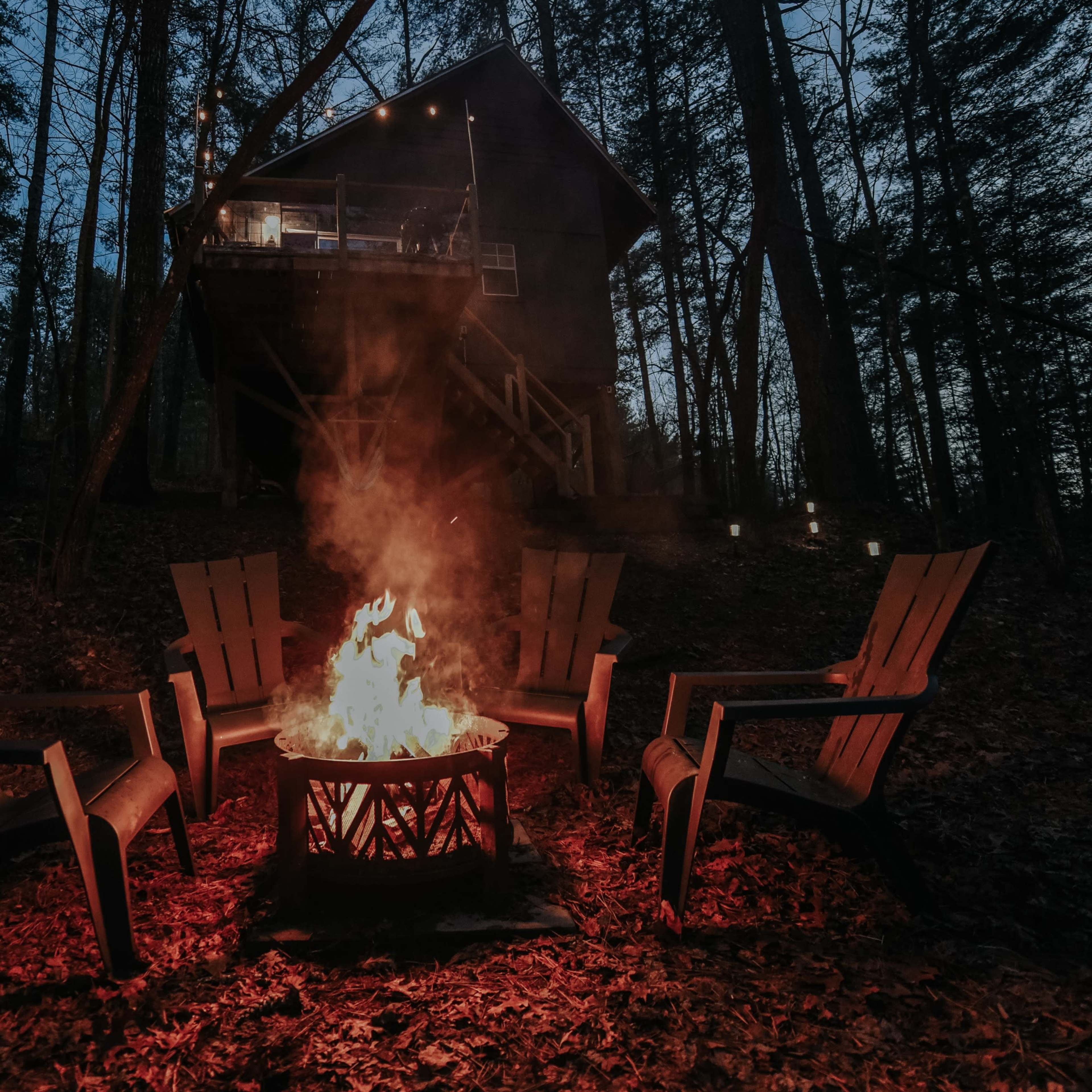 A fire pit surrounded by wooden chairs glows in front of a cabin nestled among trees at dusk.