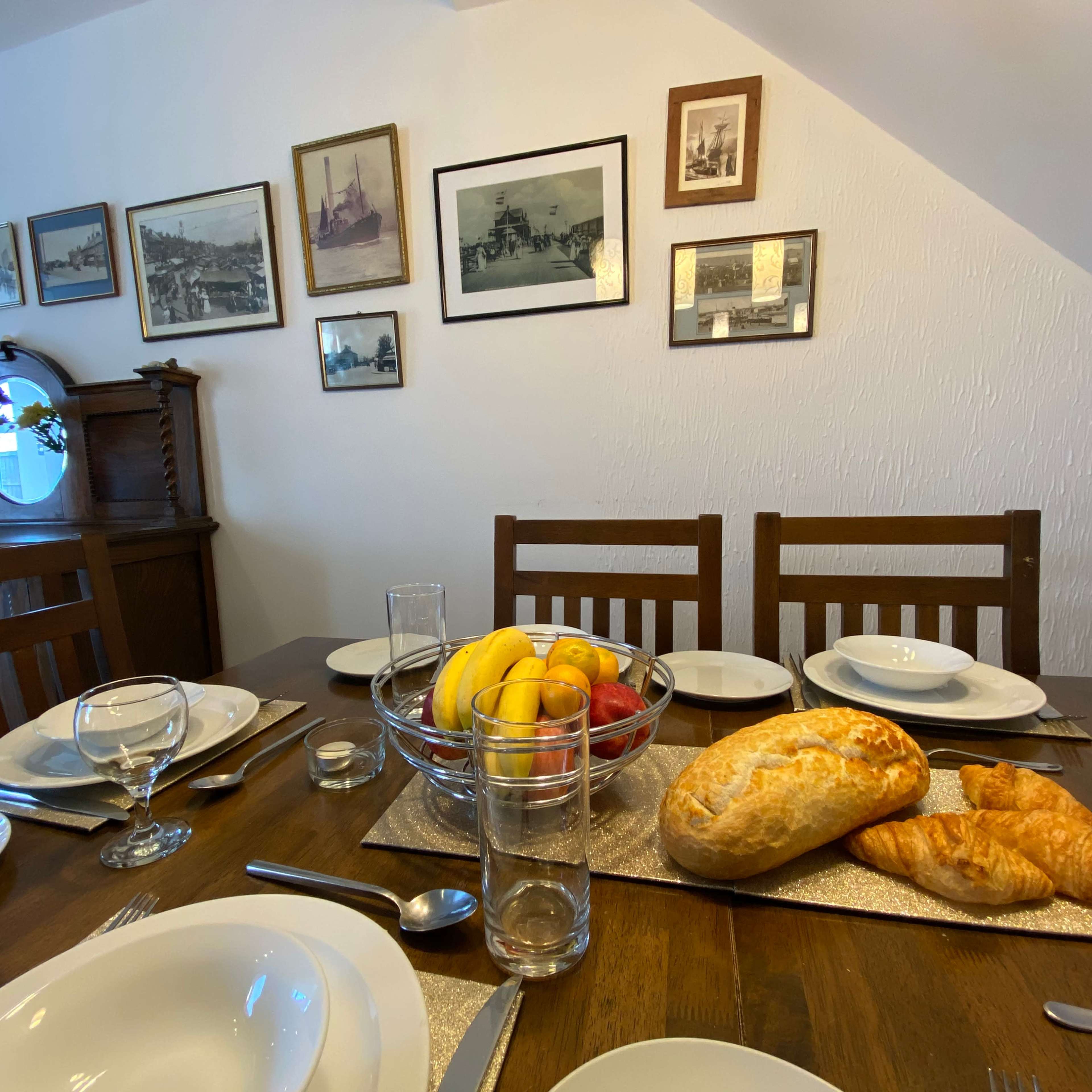 A dining table is set with plates, glasses, and a centerpiece of fruit and baked goods, while framed photographs hang on the wall behind it.
