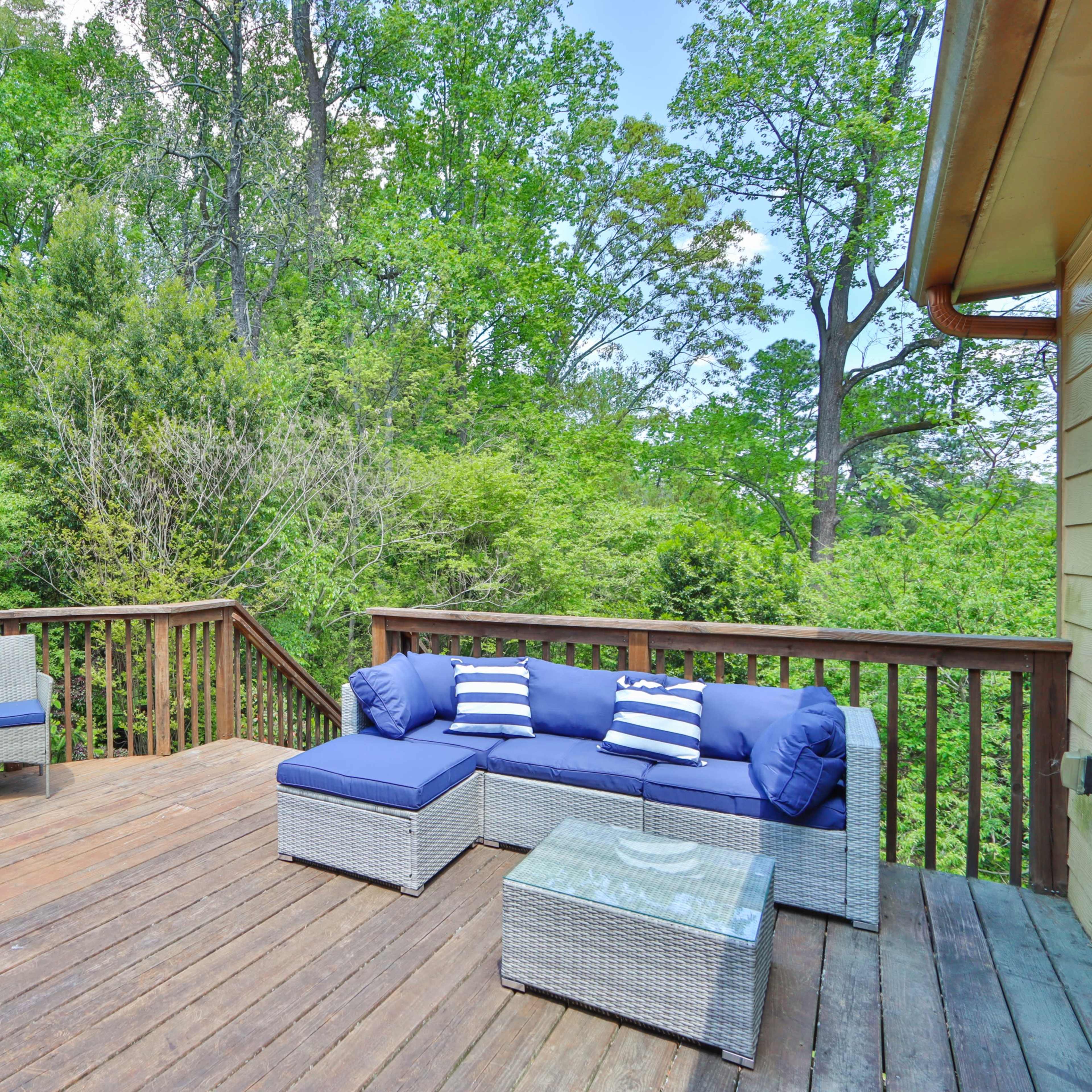 A wooden deck is furnished with a blue sectional sofa, a glass-top coffee table, and two chairs, surrounded by dense greenery.