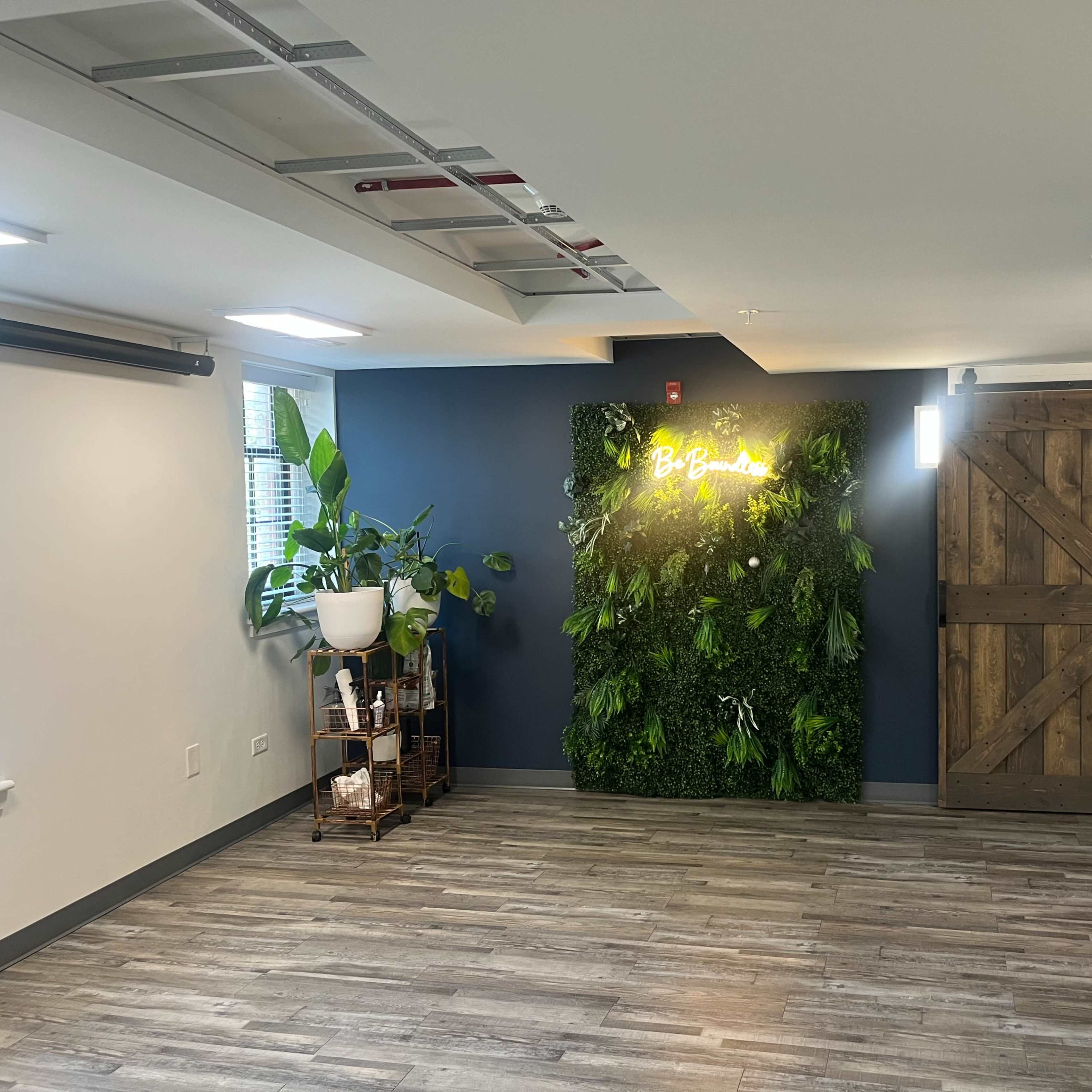 The room features a green wall with plants and a lighted sign, alongside a wooden sliding door and a small plant stand.