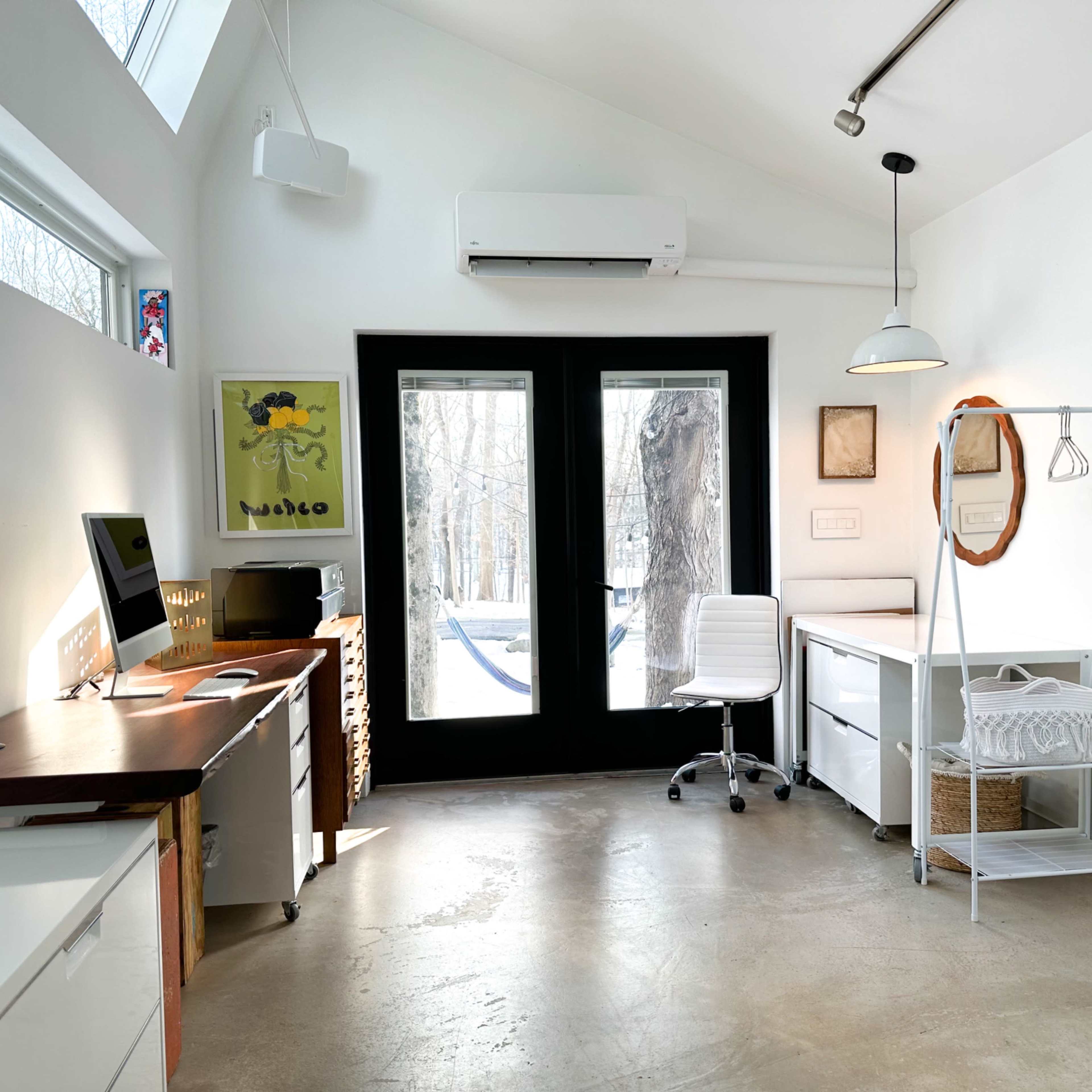 A bright, minimalist home office with large windows, a wooden desk, and a white chair, overlooking a wooded area.