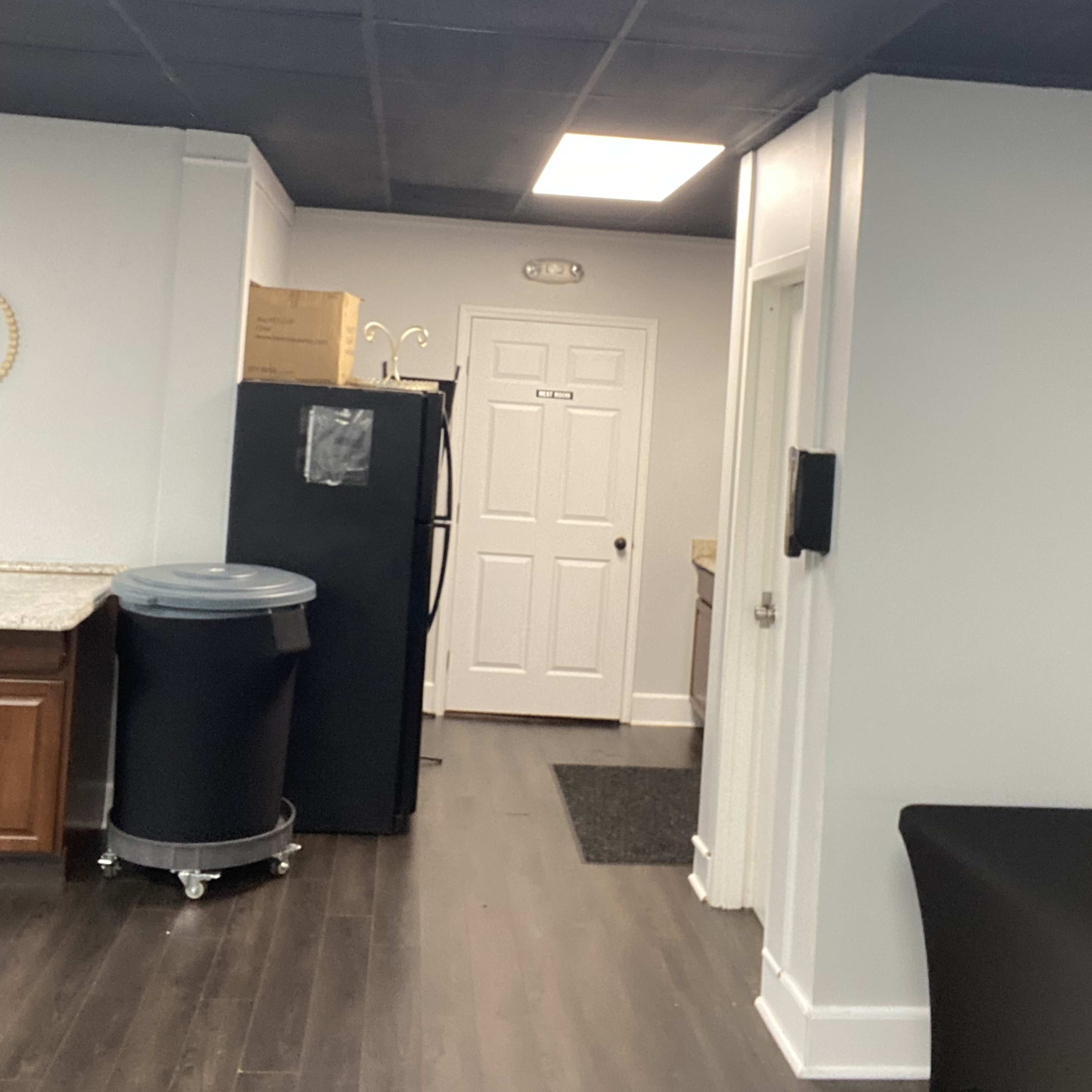 A kitchen area features a black refrigerator, a door labeled with a number, and a bright ceiling light.
