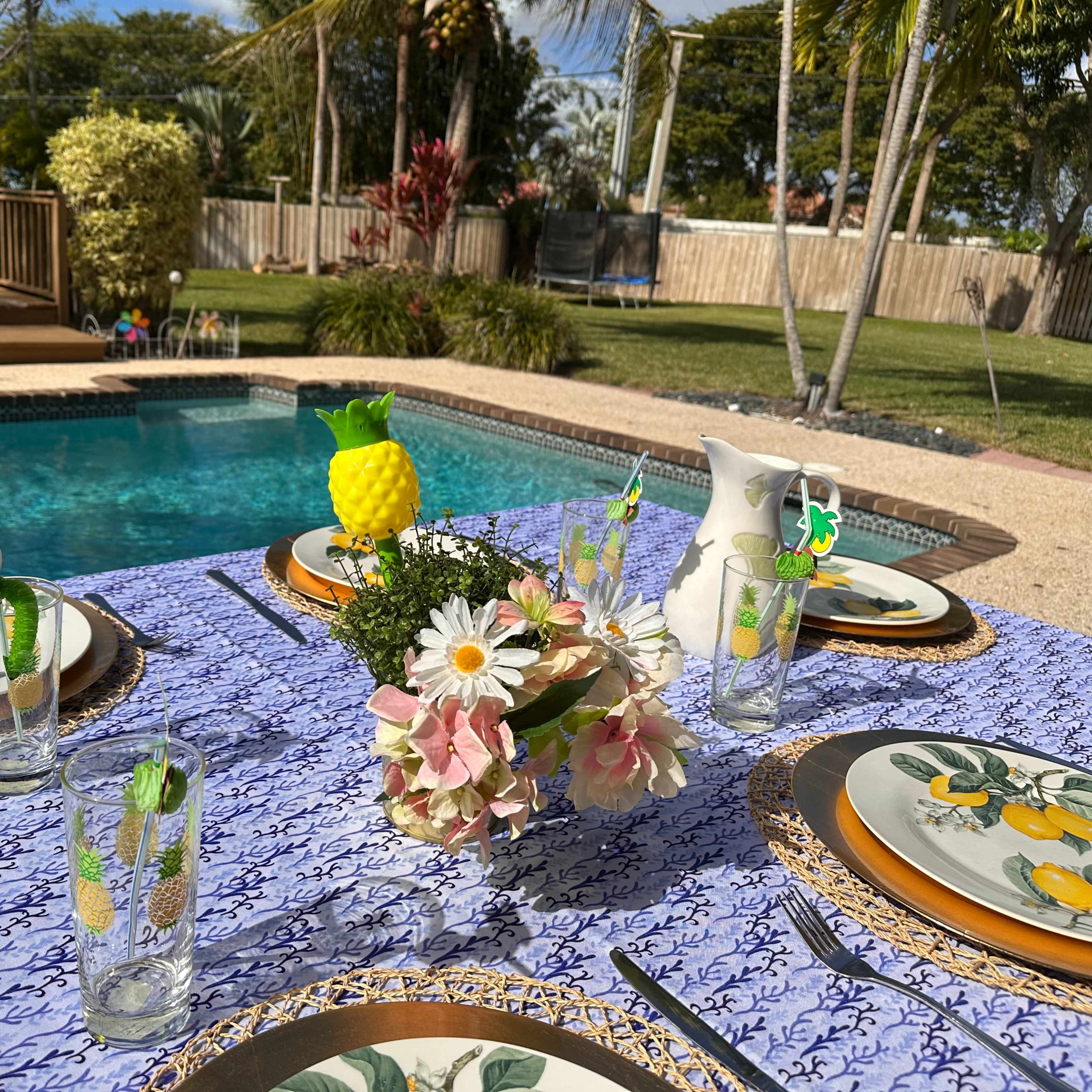 A dining table is set with decorative plates and glasses beside a swimming pool surrounded by tropical plants.