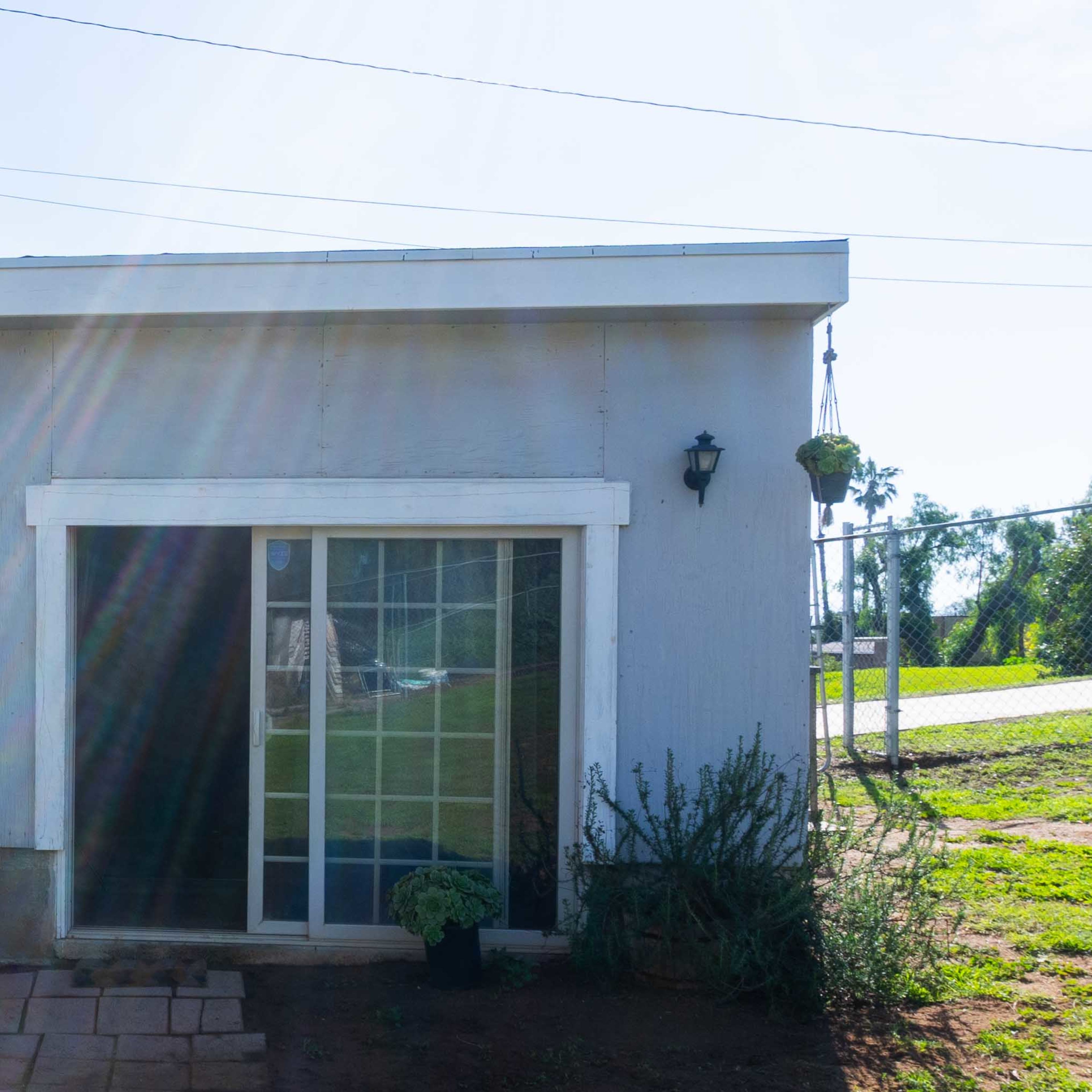 A small gray building with a sliding glass door is surrounded by plants and has two wall-mounted lights on its exterior.