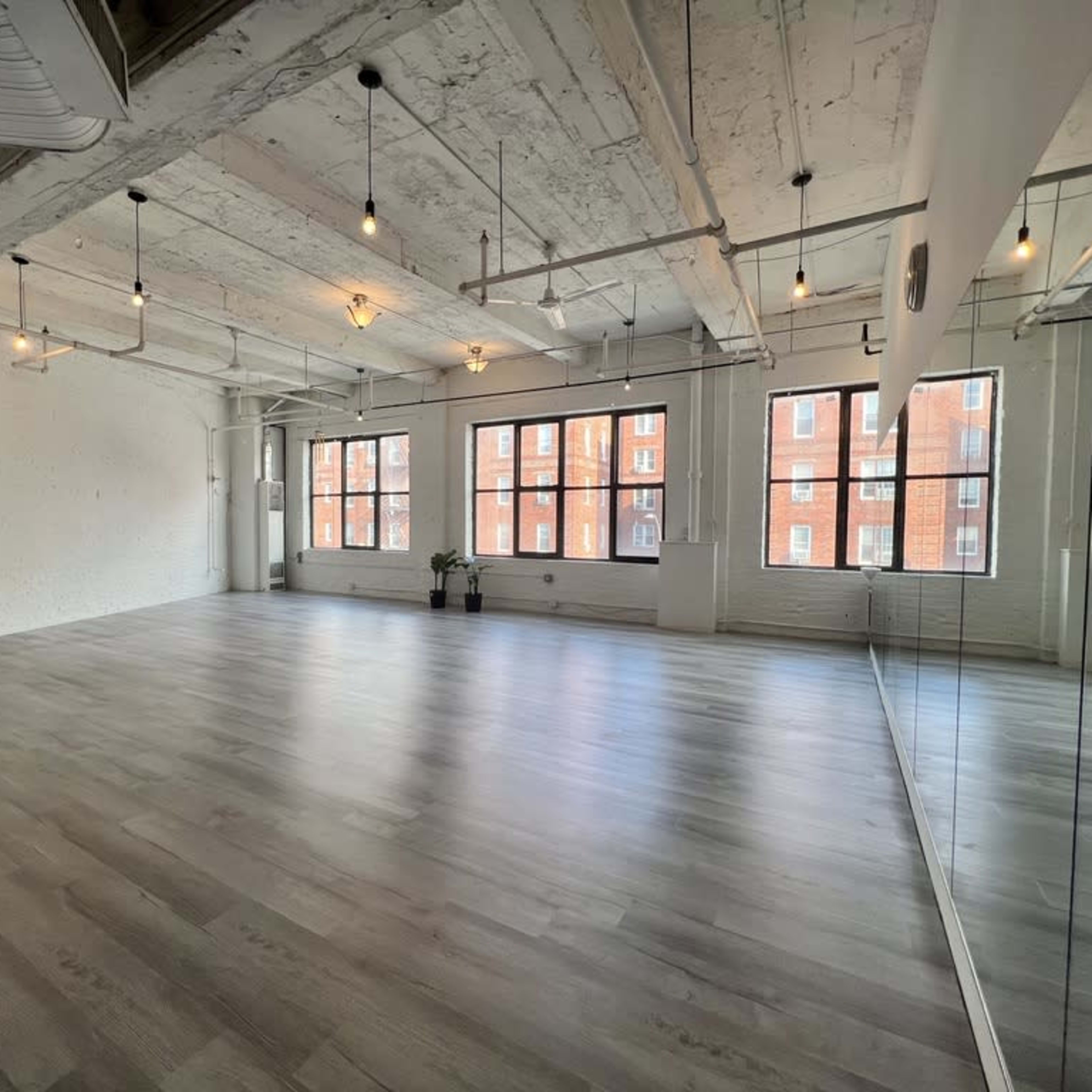 A spacious, minimalist room with large windows, wooden floors, and exposed ceilings, featuring a mirrored wall.