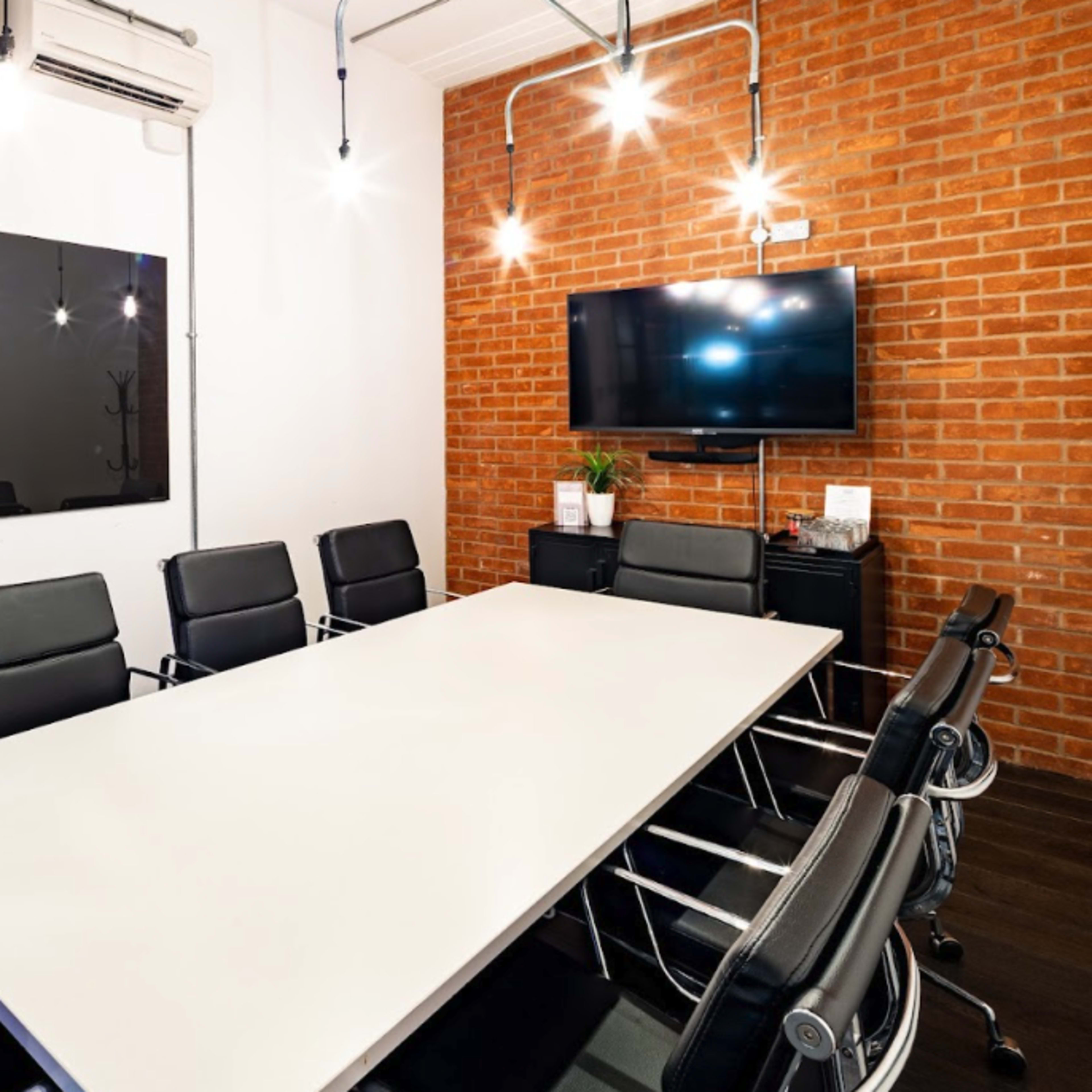 A modern conference room features a long white table surrounded by black chairs, with a brick accent wall and two screens mounted on opposite walls.