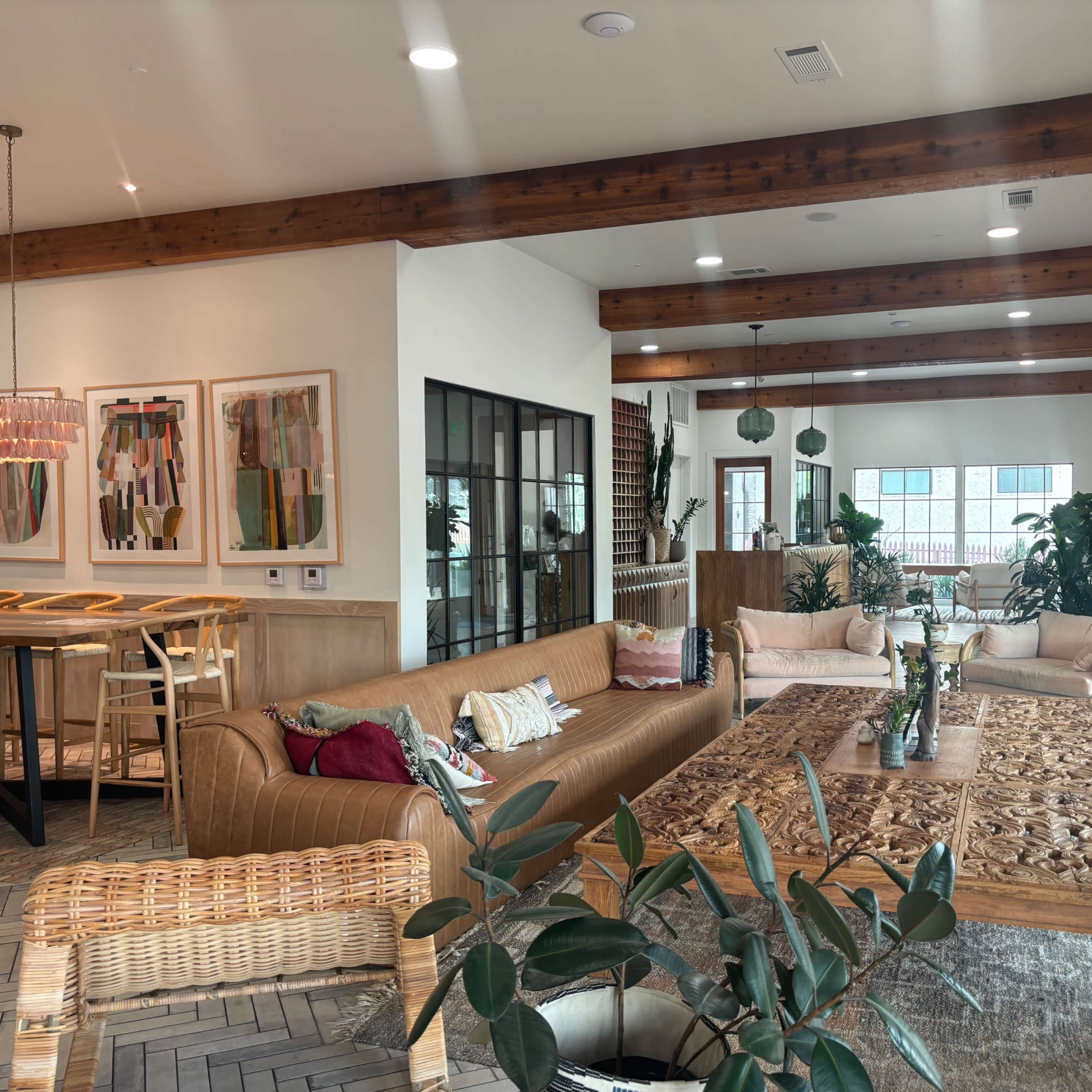 A modern living area with a large wooden coffee table, a brown leather sofa, dining area, and plants throughout the space.