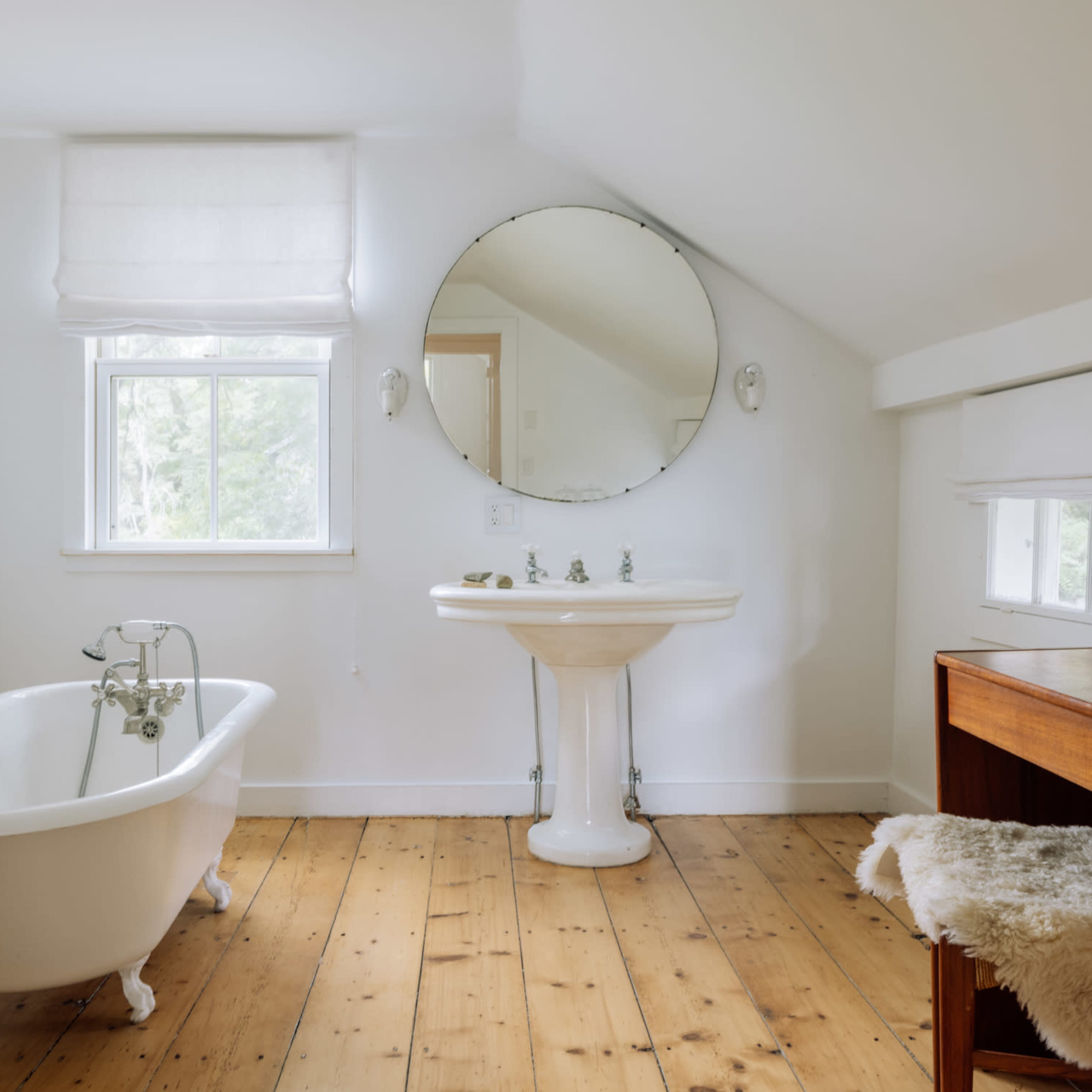 A bright bathroom features a freestanding bathtub, a pedestal sink, and a wooden desk with a fur seat on a wooden floor.
