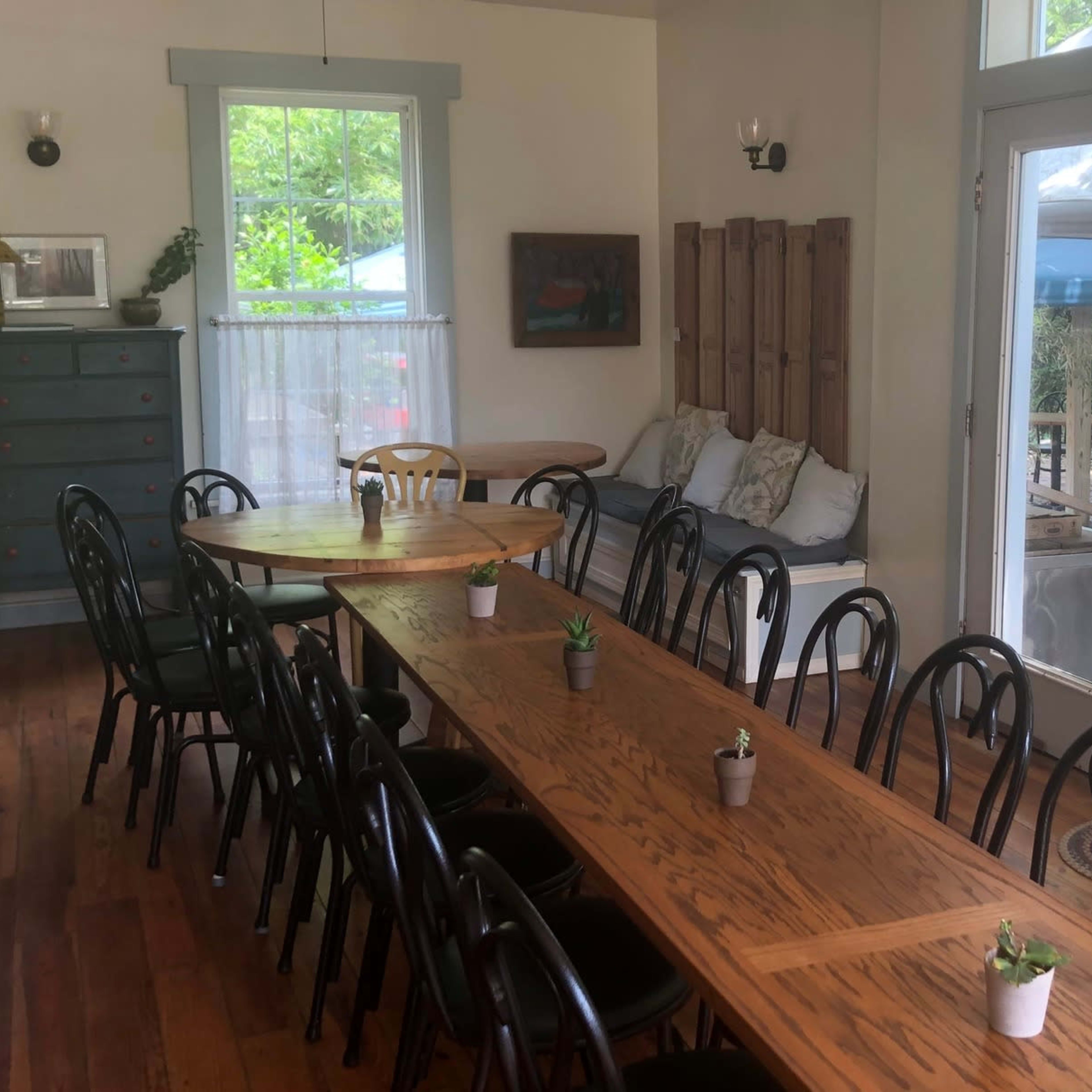 A dining area with a long wooden table surrounded by black chairs, large windows letting in natural light, and a door leading to an outdoor space with seating.