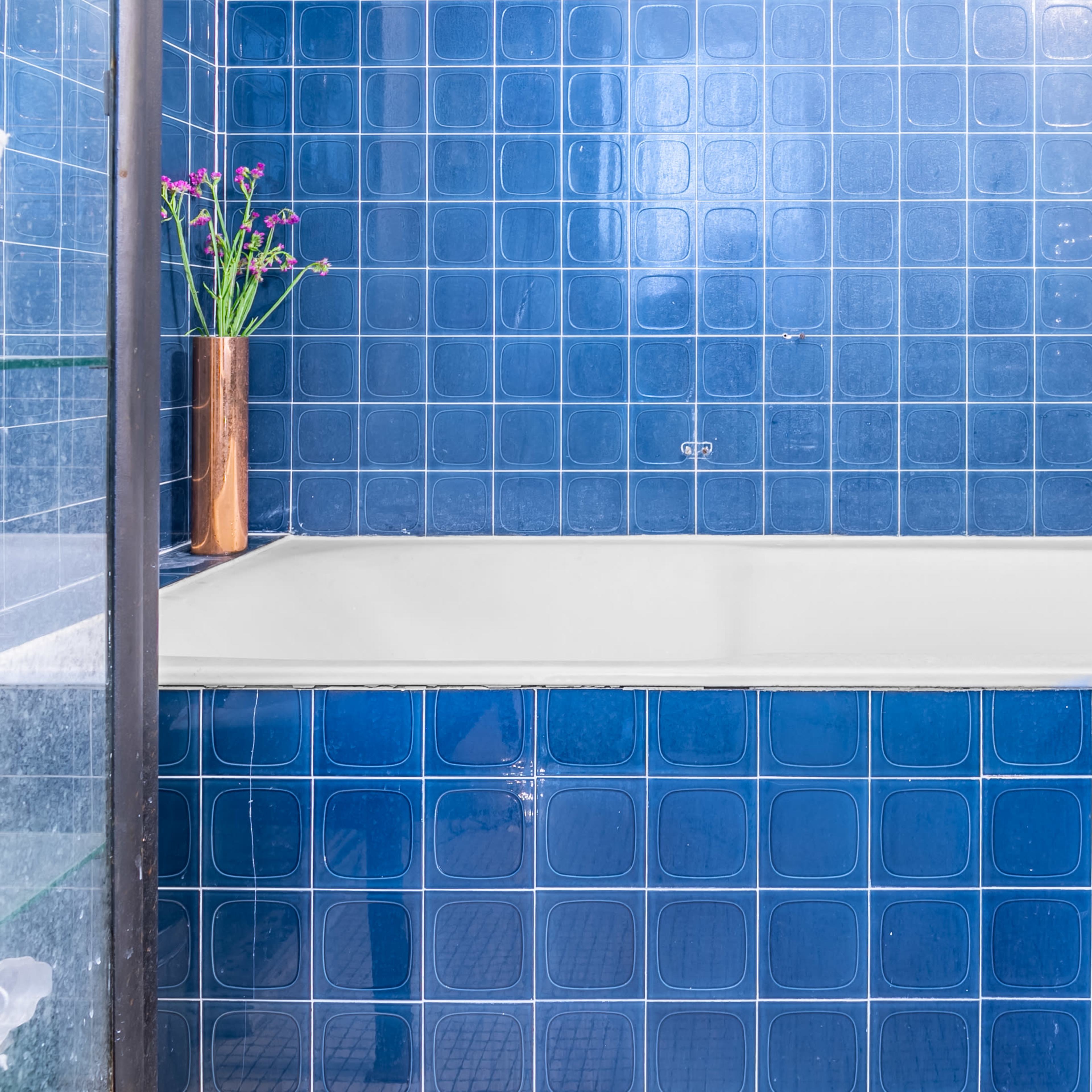 A blue-tiled bathroom features a white bathtub and a small vase with flowers on the edge.