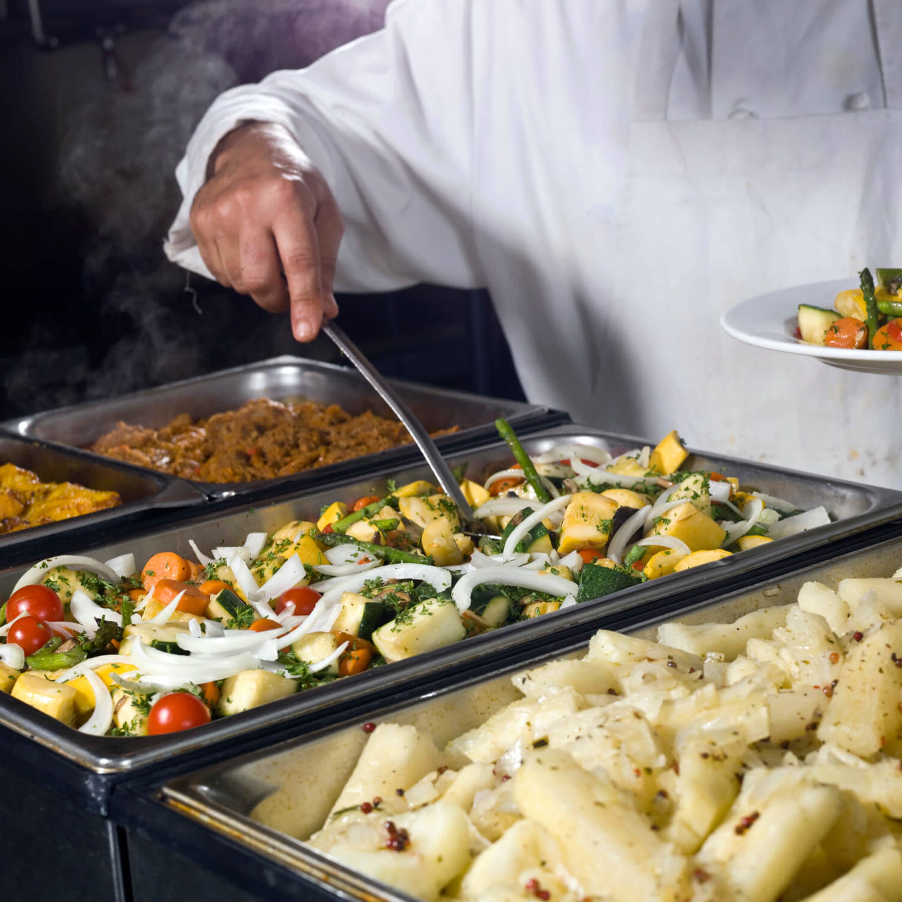 A chef serves a plate of mixed vegetables from a buffet line filled with various dishes.