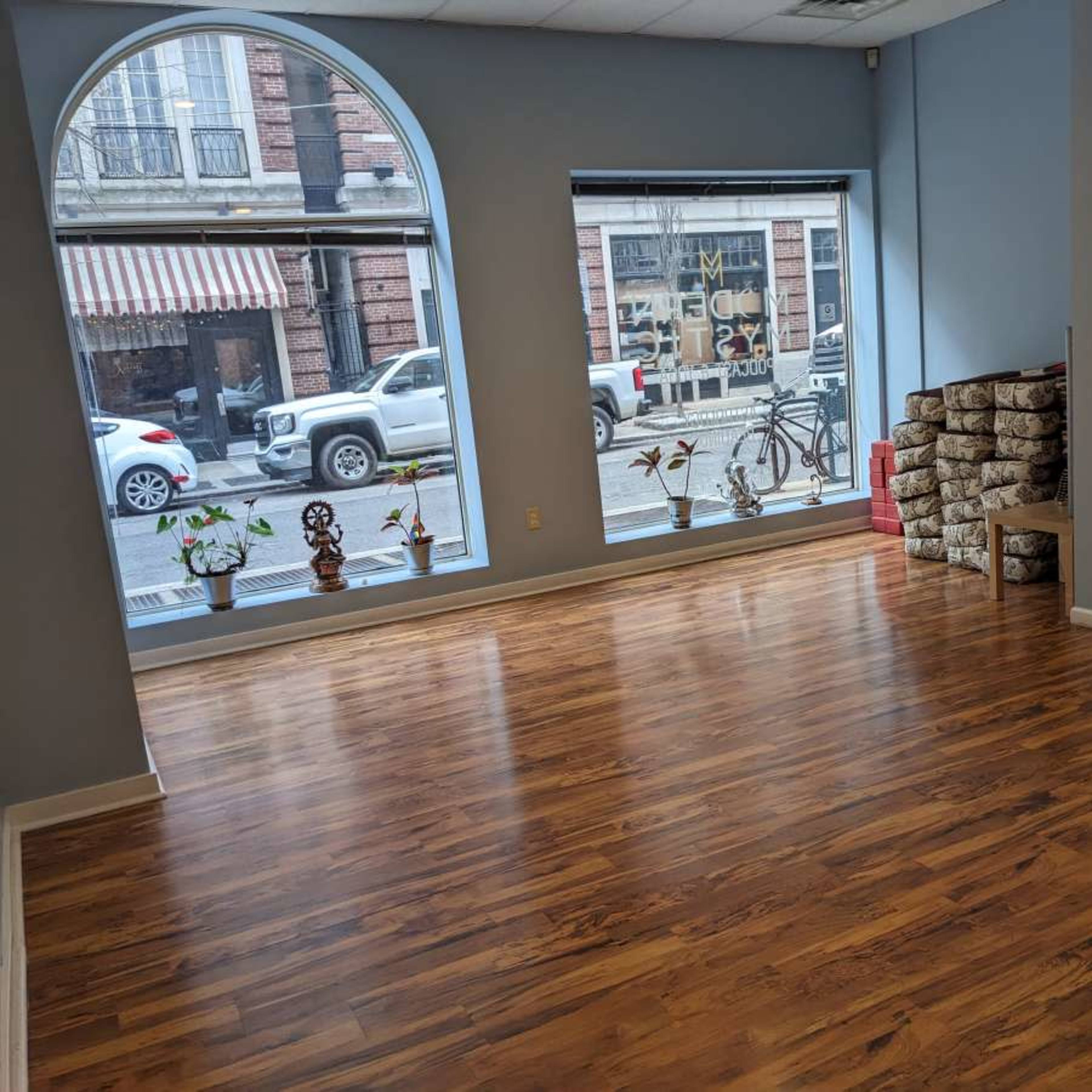 A spacious, empty room with wooden flooring and large windows displaying a street view.