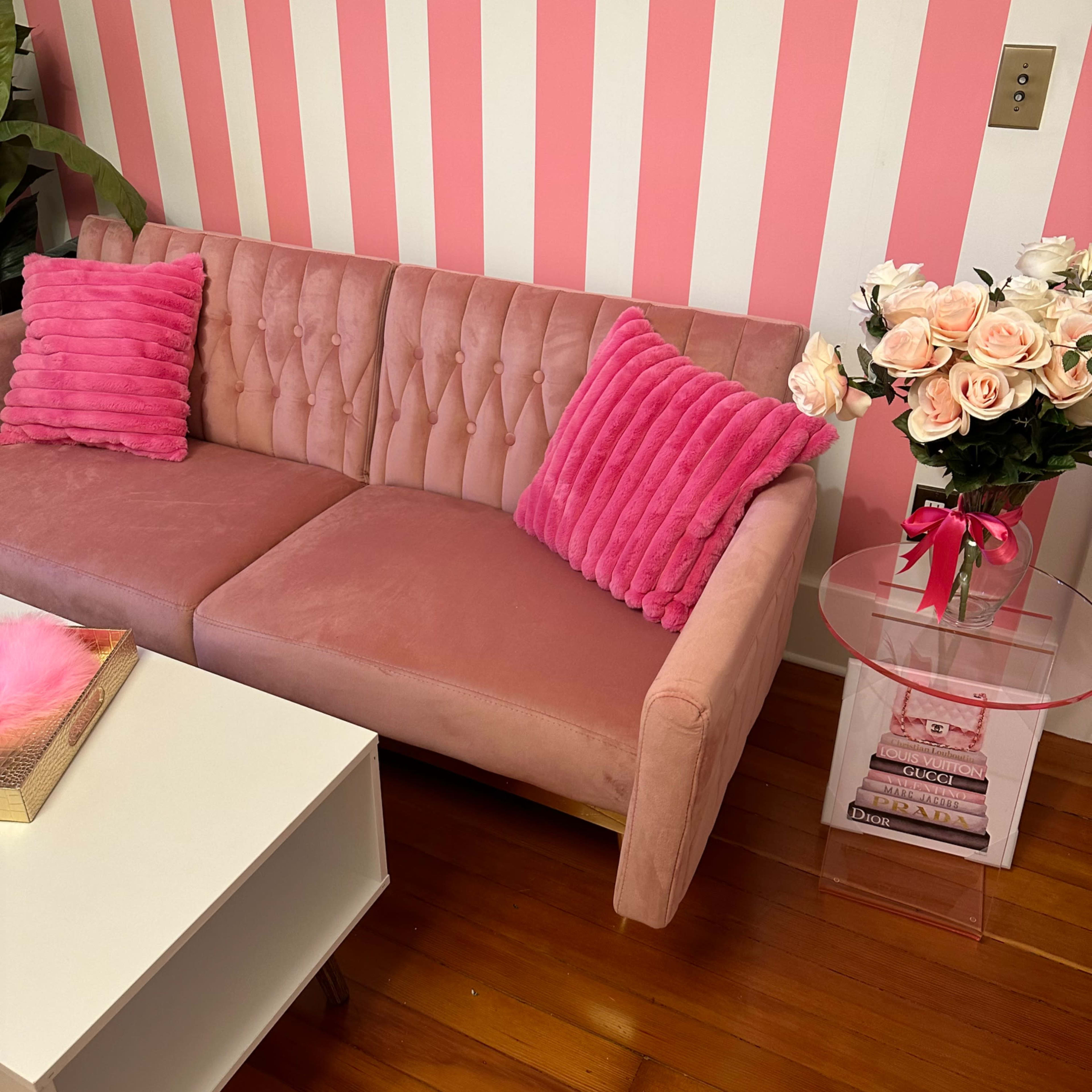 A pink tufted couch with matching cushions sits beside a clear side table adorned with a vase of roses and a small stack of books, against a striped pink wall.