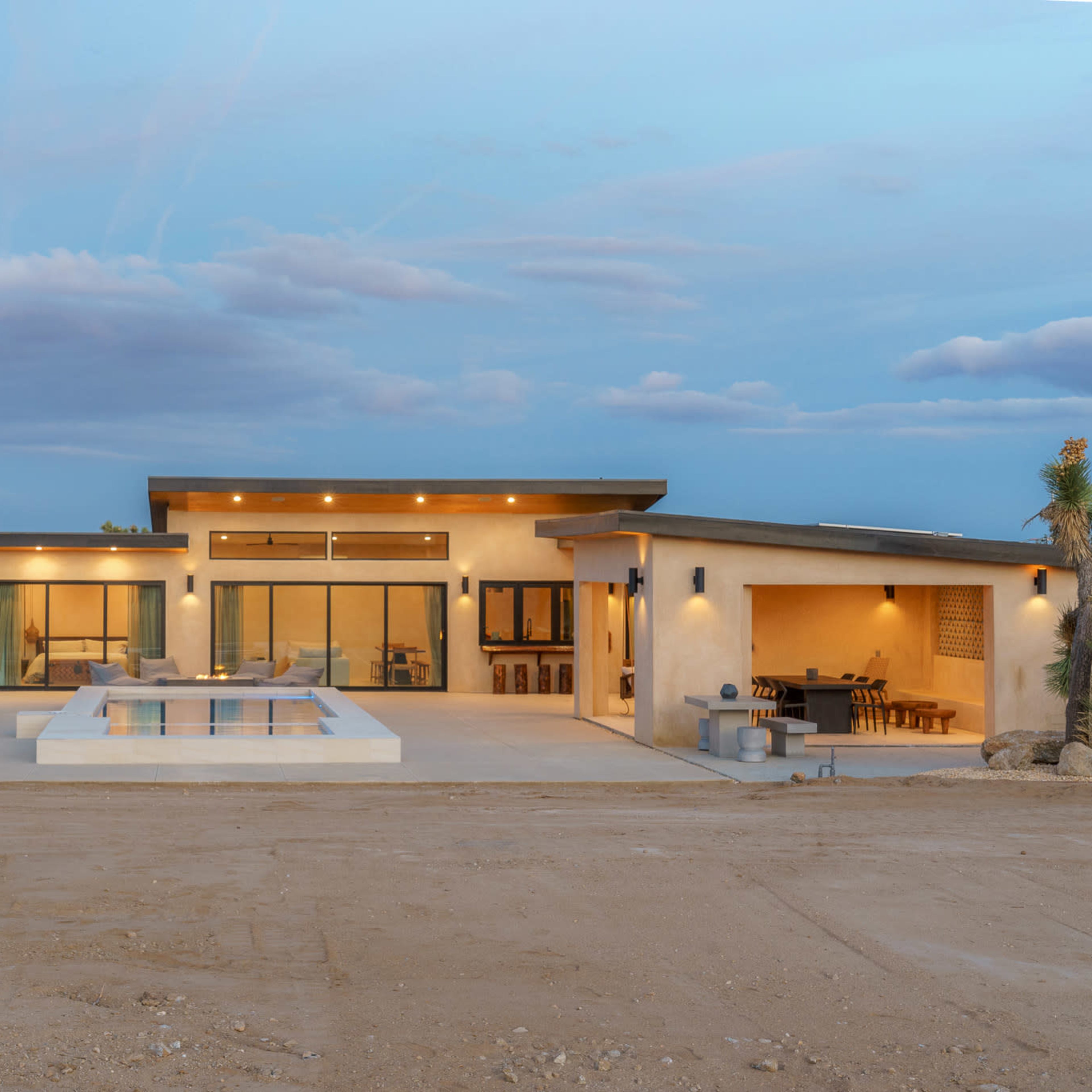 A modern single-story house with a pool, large windows, and a desert landscape featuring a Joshua tree in the foreground.