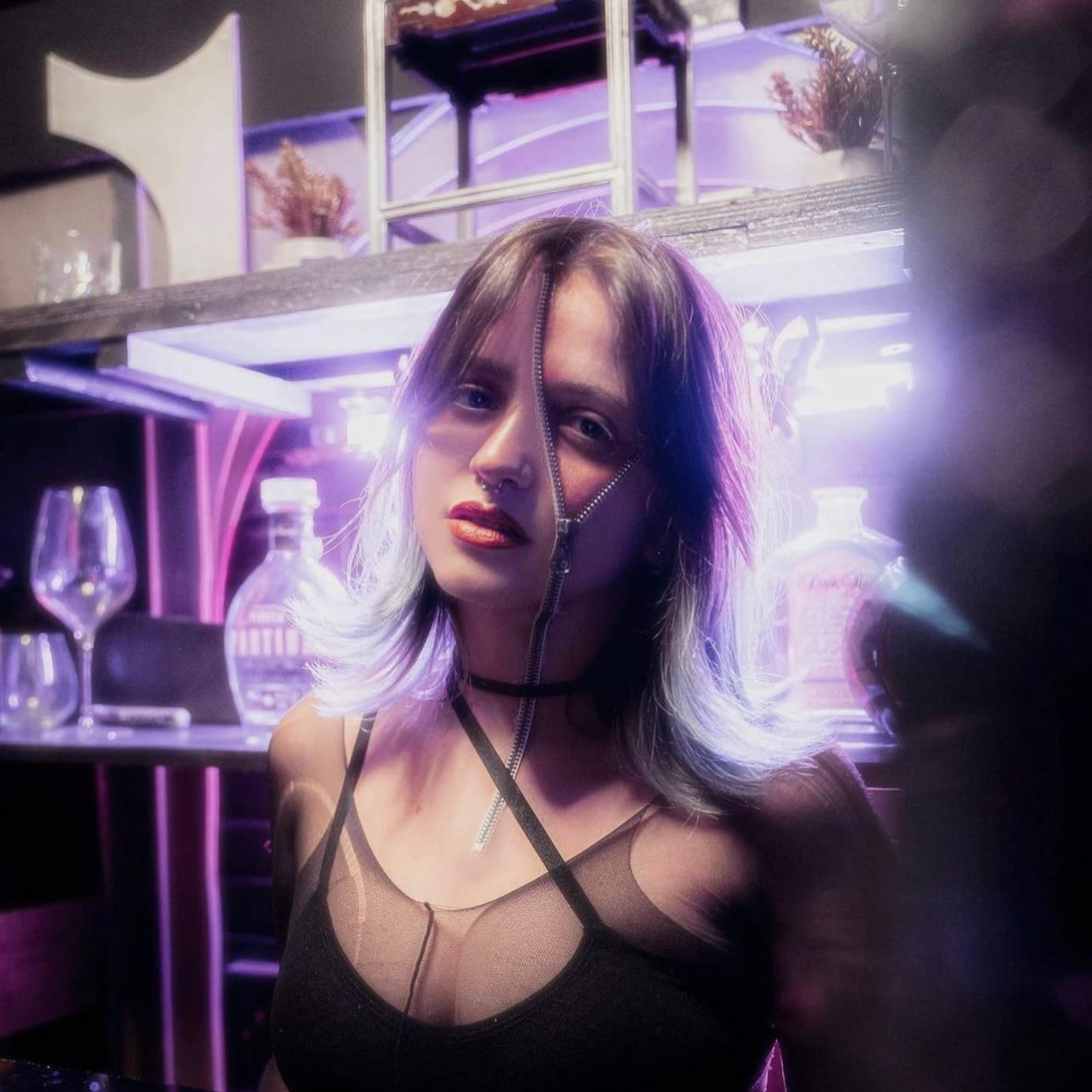 A woman with purple-tinted hair is standing in front of a bar with illuminated shelves and glassware.