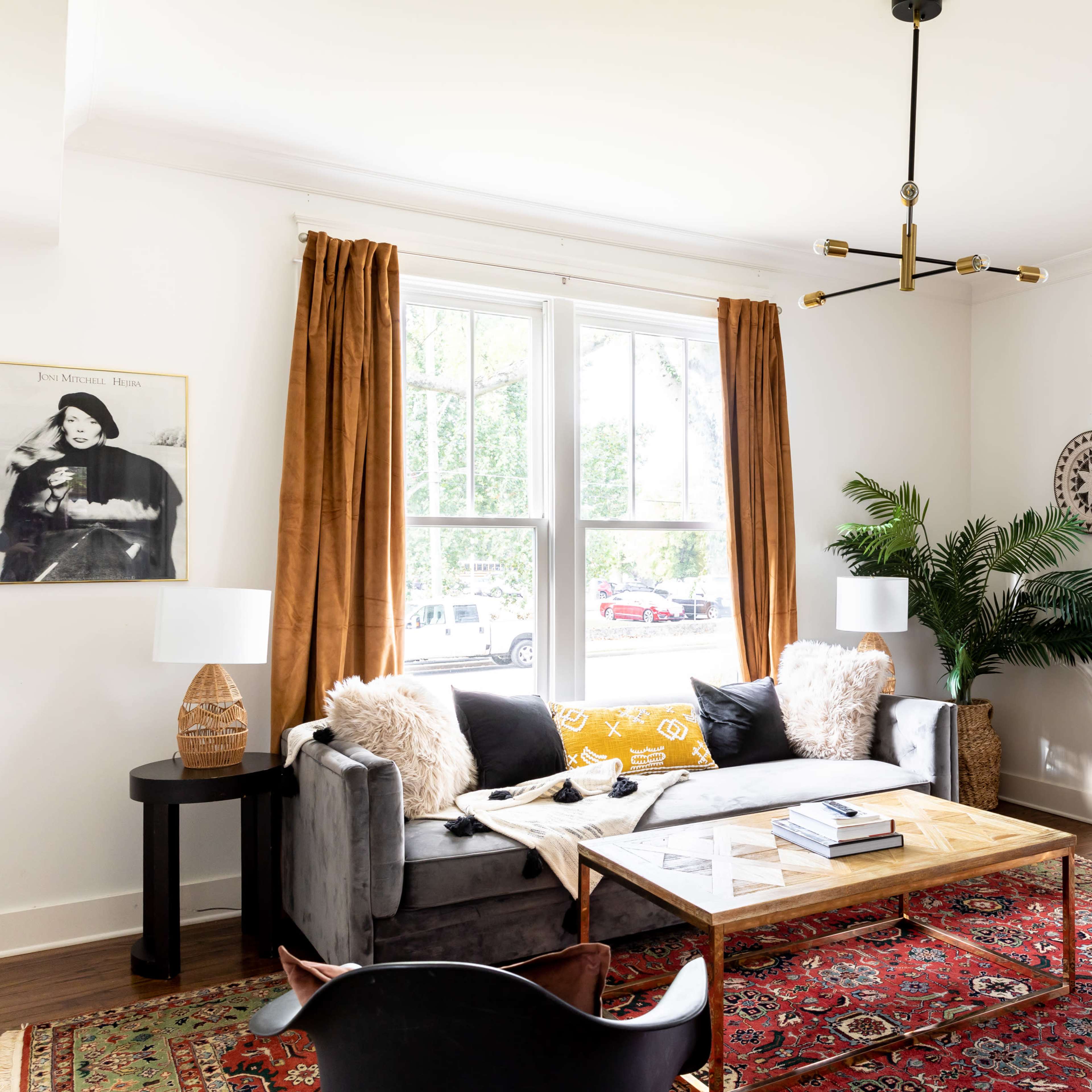 A living room features a gray sofa with decorative pillows, a wooden coffee table, and a mix of plants and artwork along the walls.