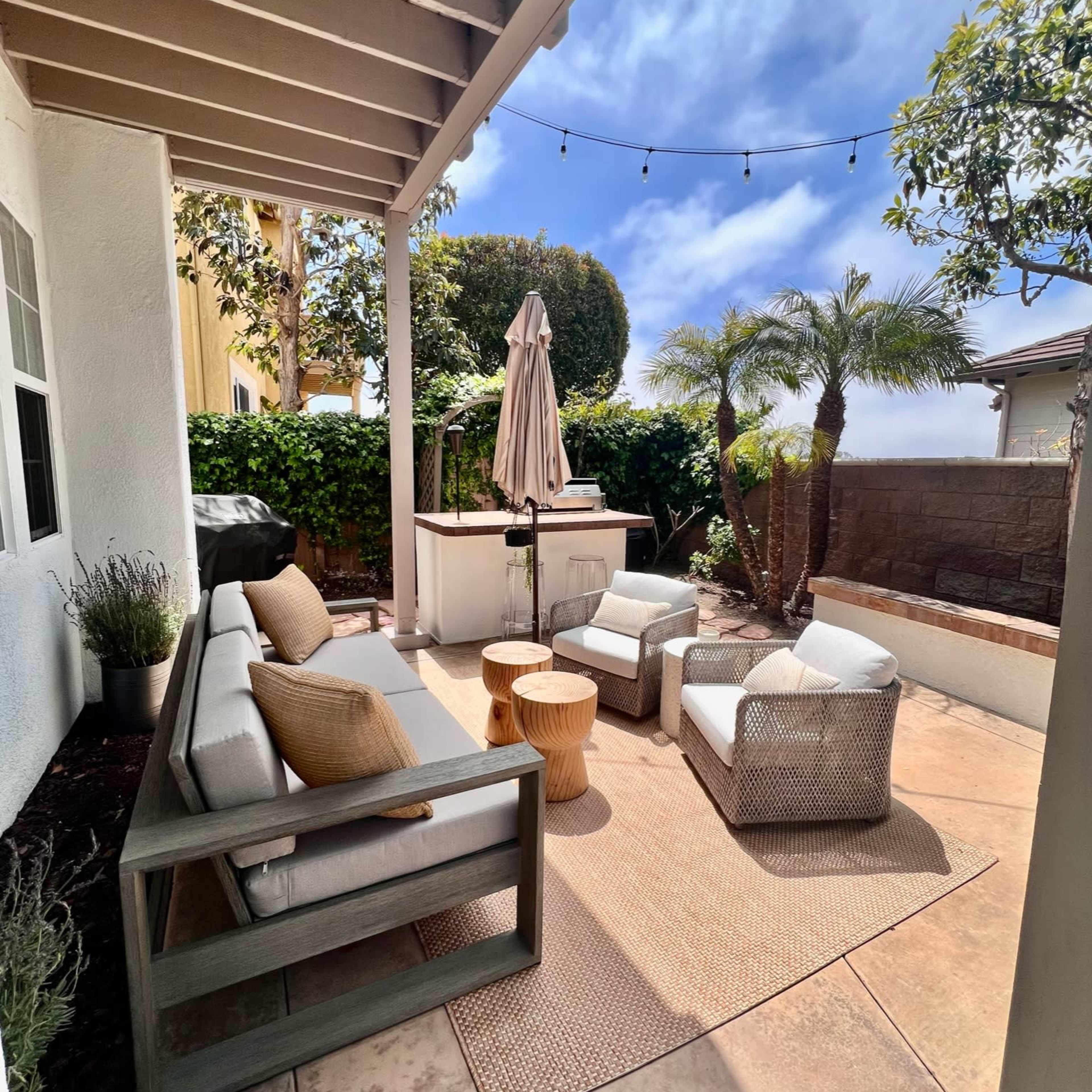 A patio area with a sectional sofa, a small wooden table, and two lounge chairs surrounded by plants, with a grill and an umbrella visible in the background.
