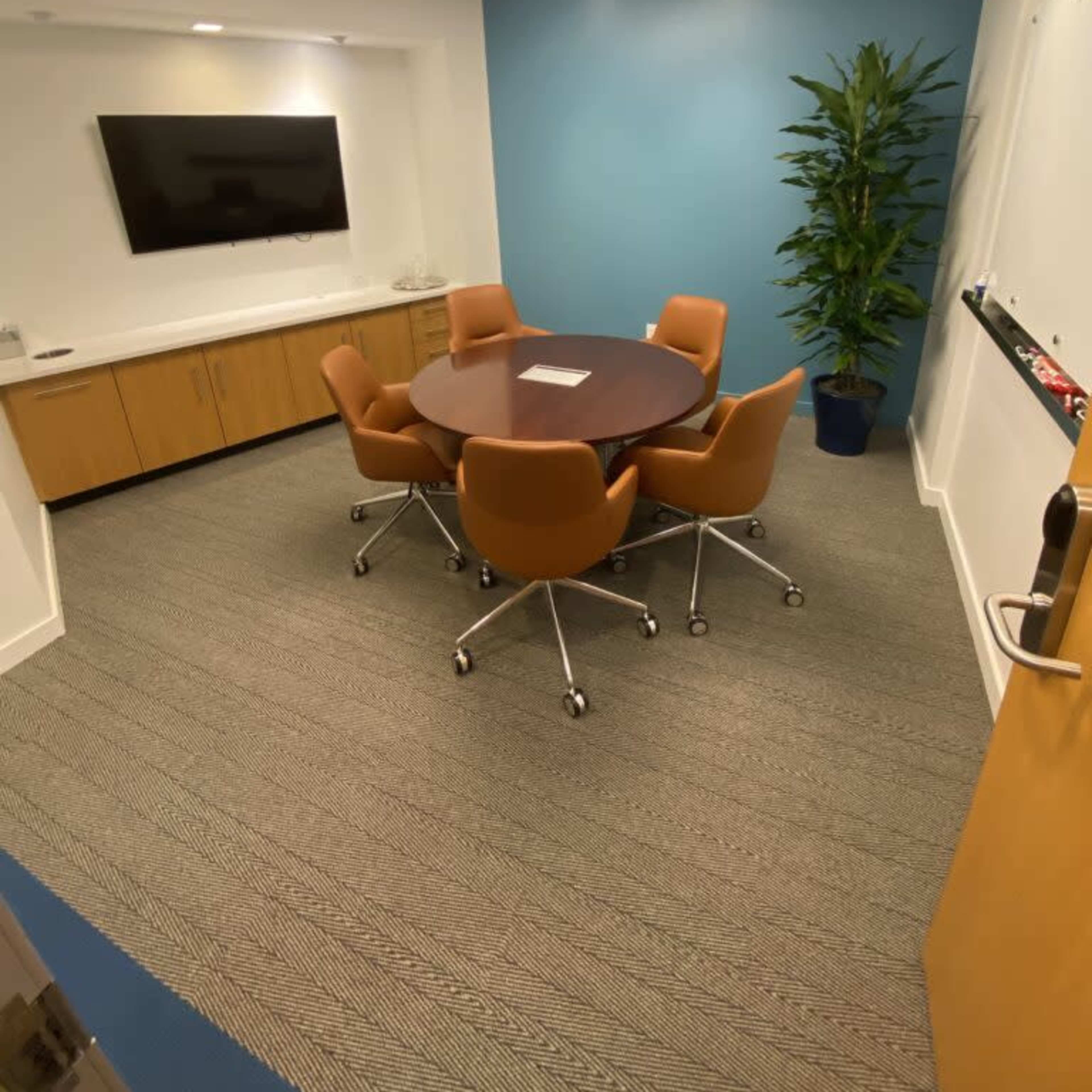 A small conference room with a round table, six chairs, a large wall-mounted television, and a potted plant in the corner.
