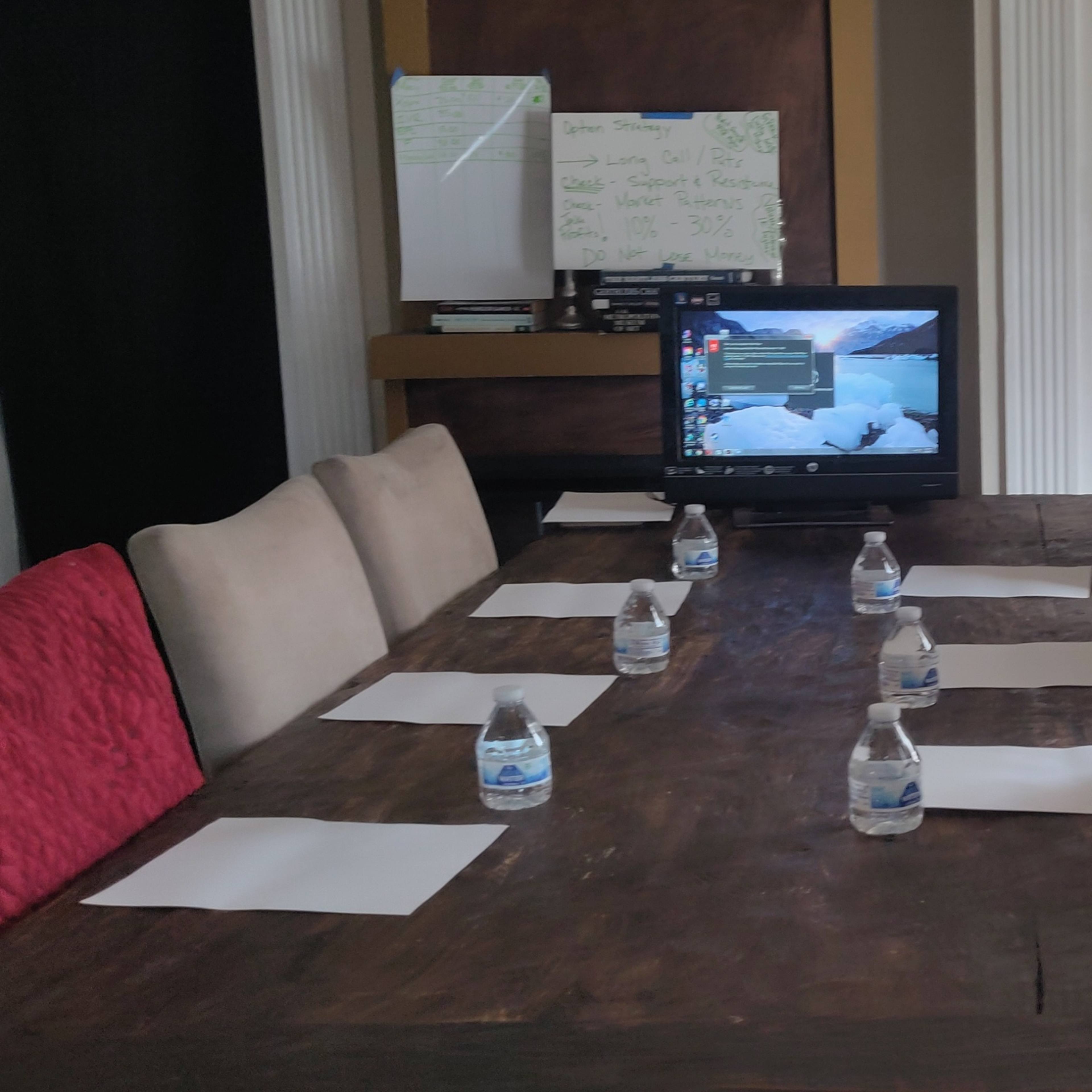 A long wooden dining table is set with bottles of water and blank papers, while a television displaying a menu screen is positioned at one end of the room.