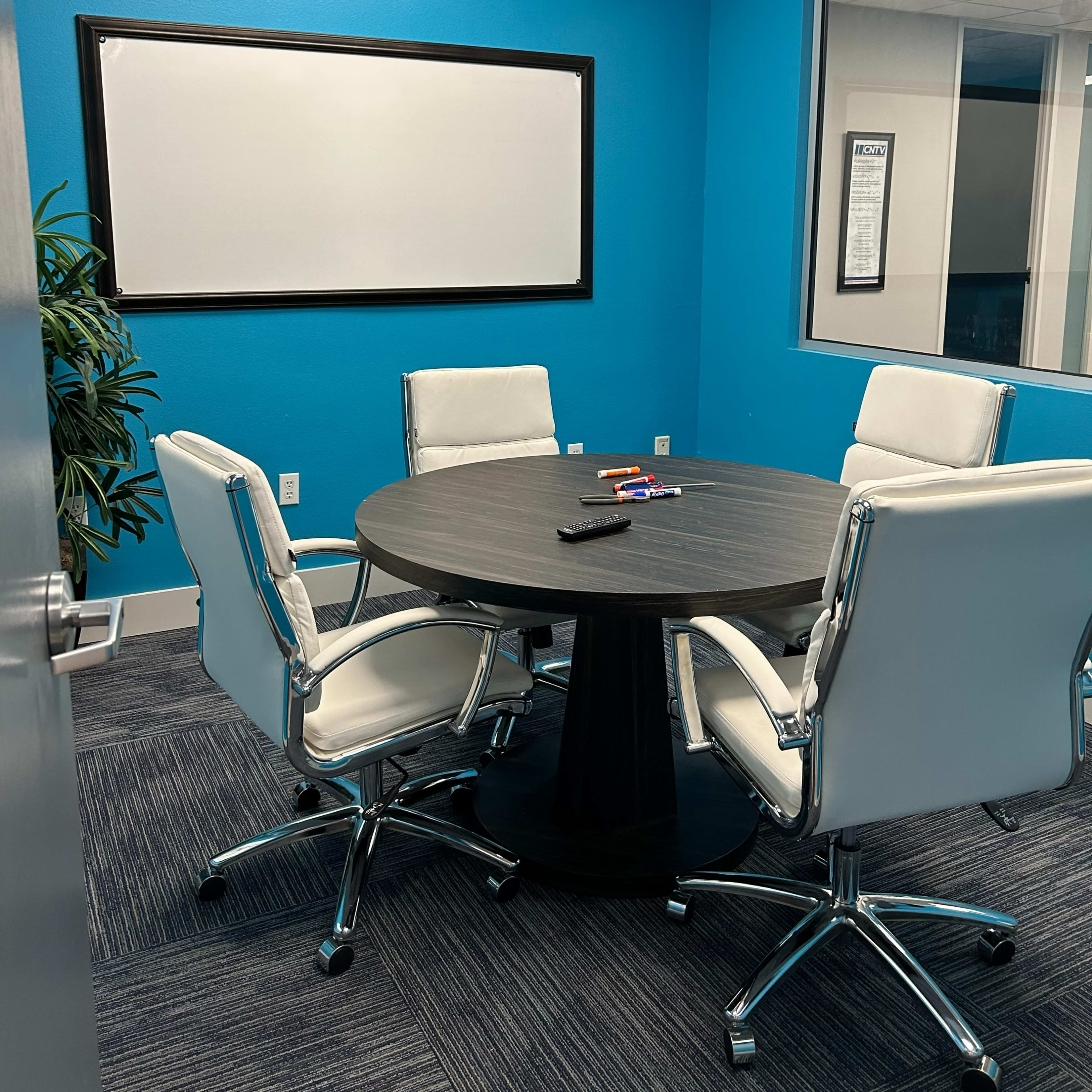 A small conference room with a round black table and four white chairs, set against a bright blue wall and featuring a whiteboard.