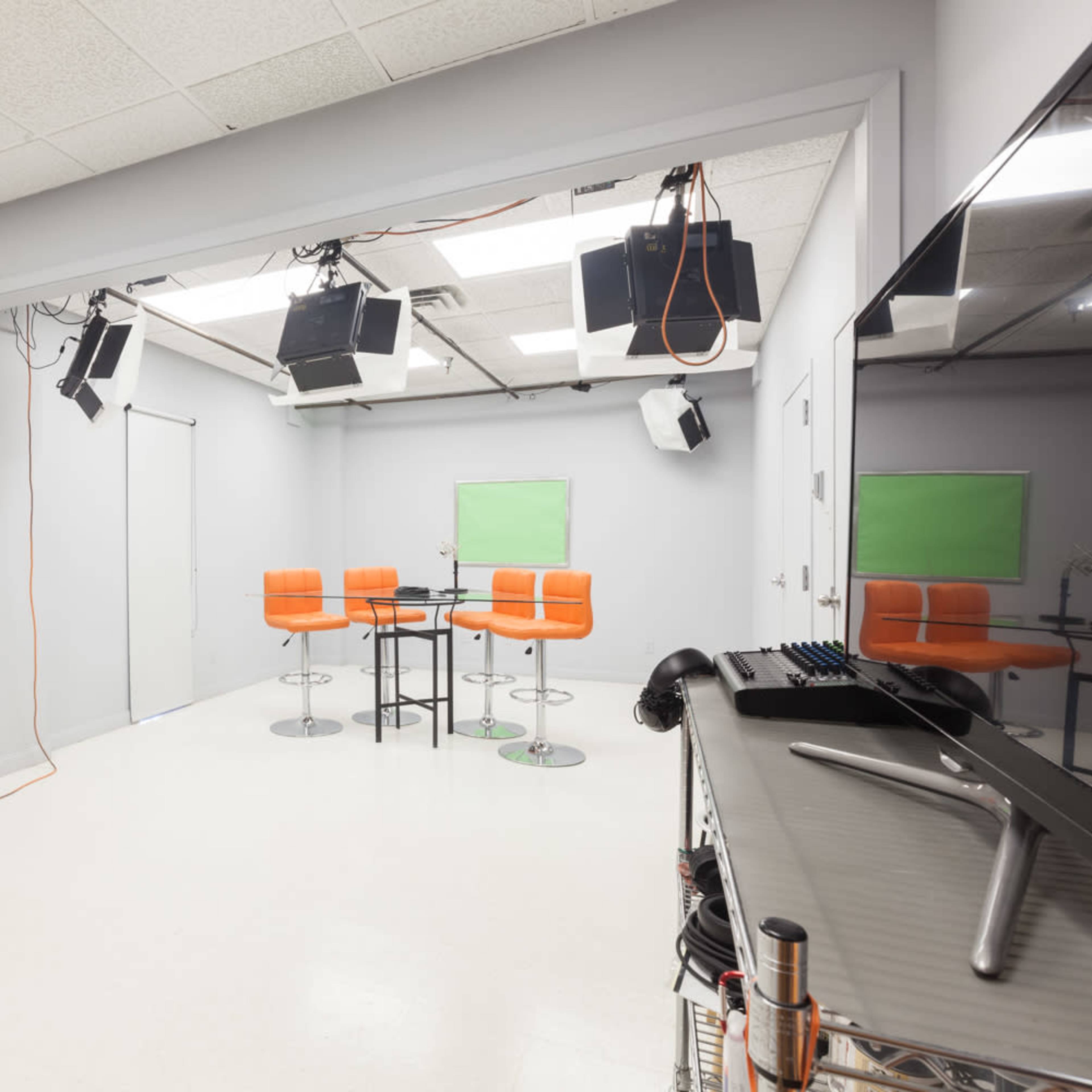 A modern studio space features orange bar stools around a black table, with professional lighting equipment and green screen areas.