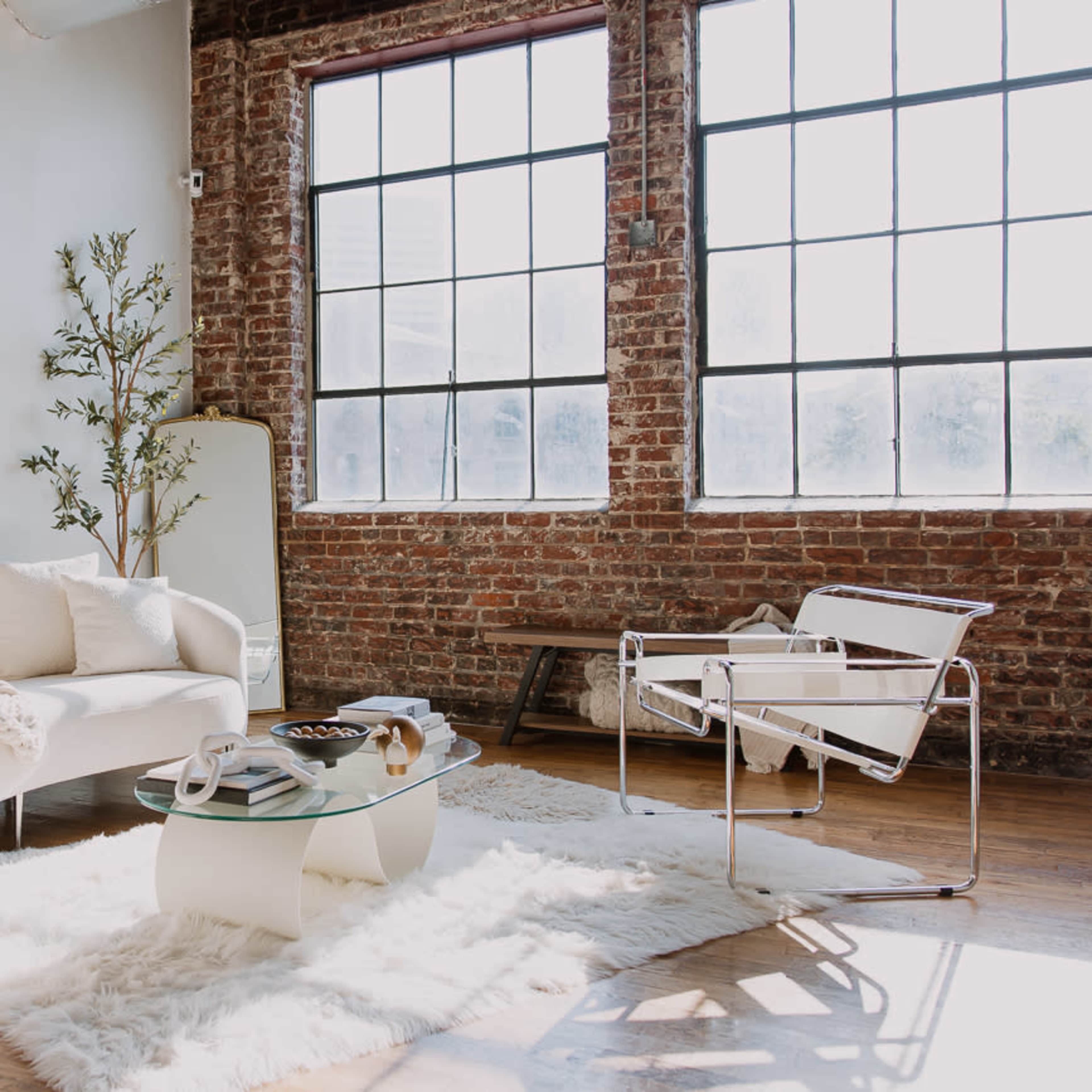 A bright living space features a white sofa and chair set, a glass coffee table, and large windows framed by exposed brick walls.
