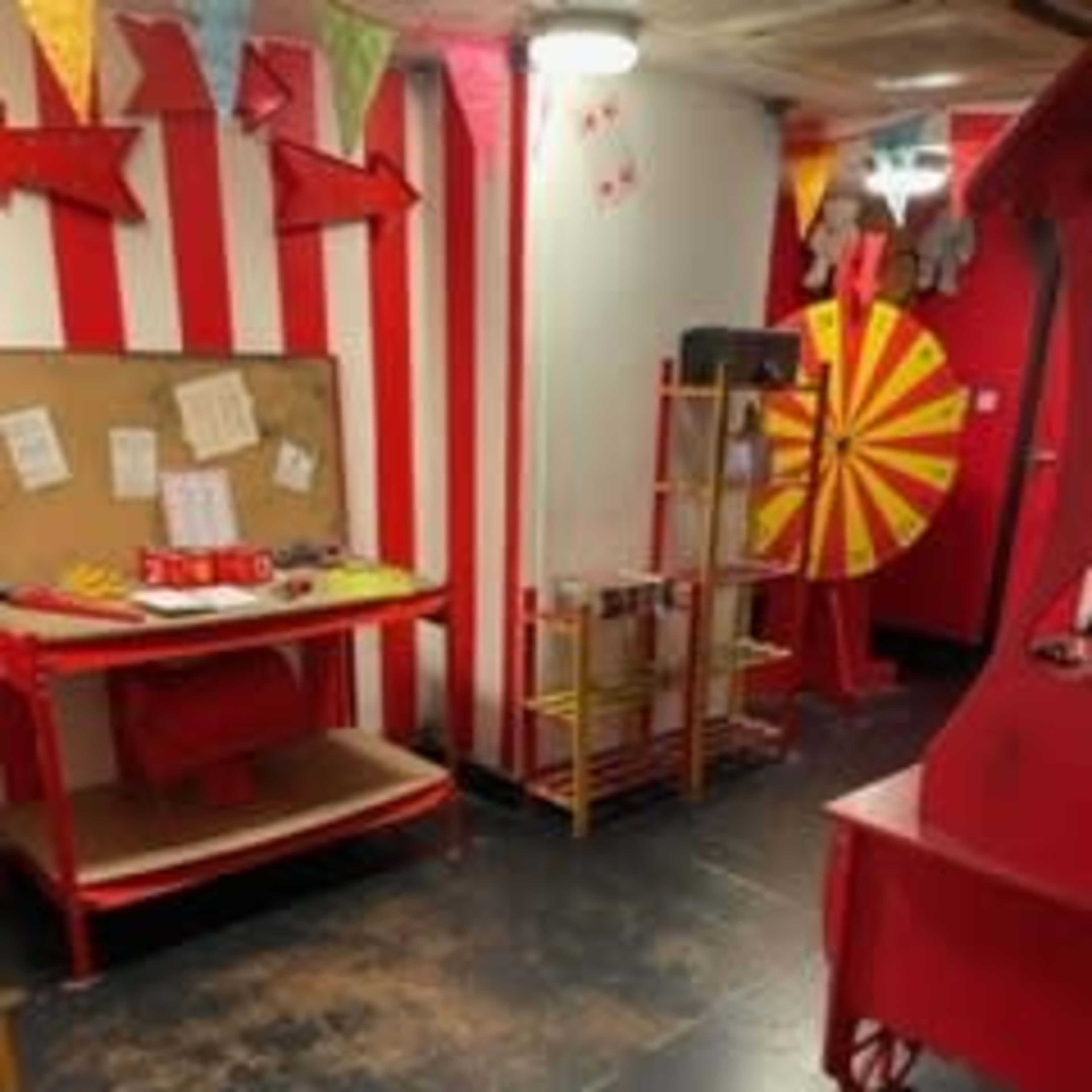 The scene shows a carnival-themed room with red and white striped walls, a bulletin board, a spinning prize wheel, and various festive decorations.