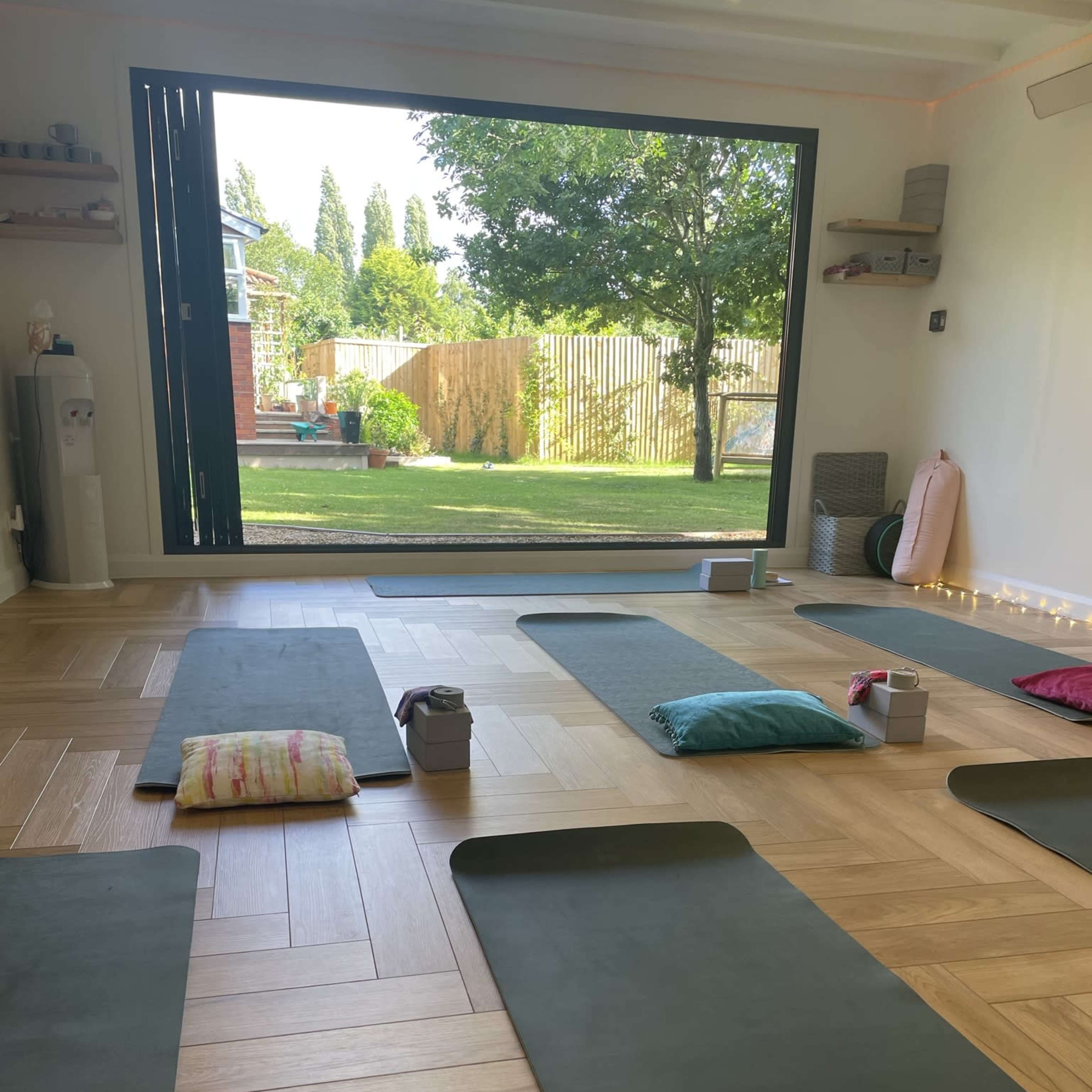 A yoga studio with green mats arranged on a wooden floor, overlooking a garden through a large window.