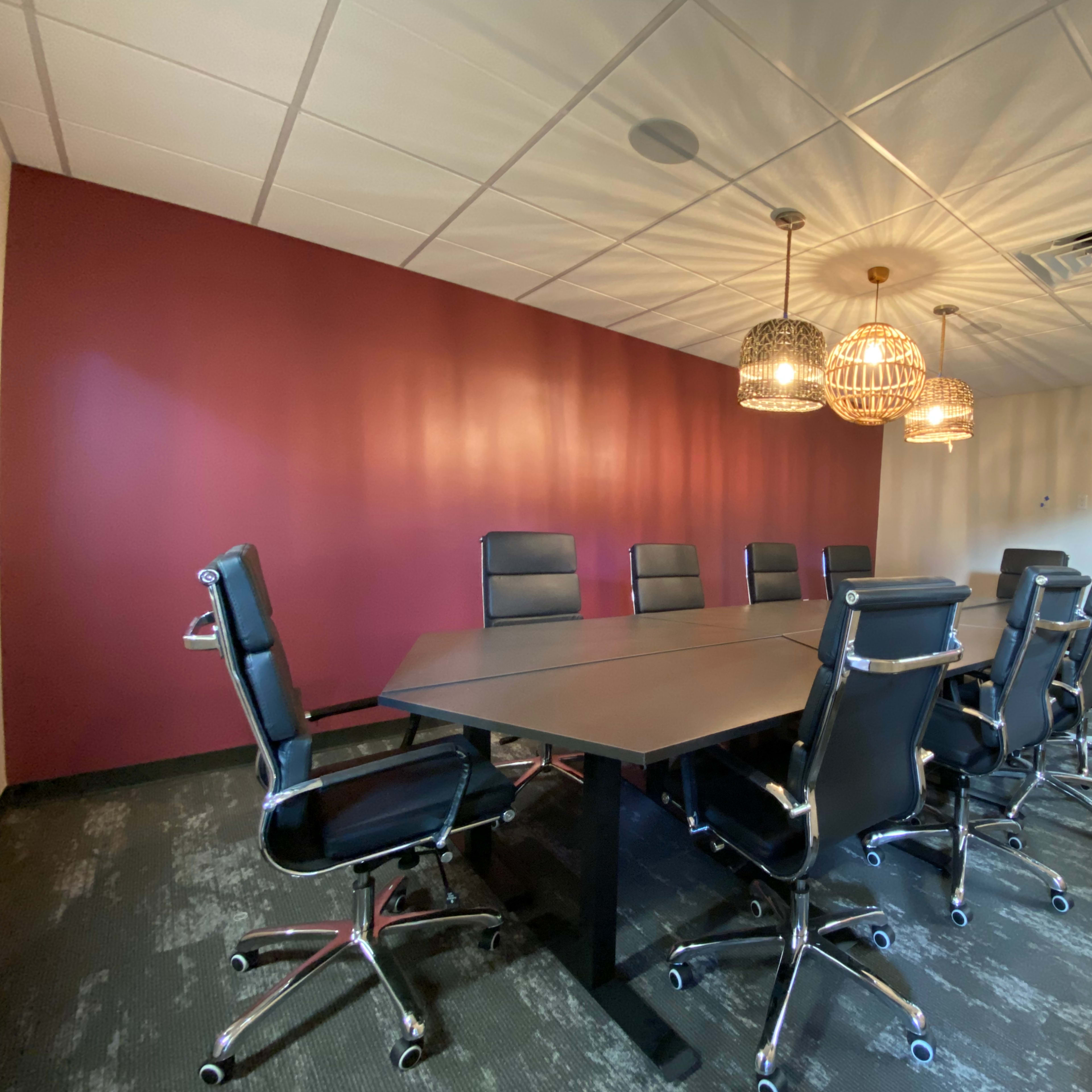 A conference room features a long, dark table surrounded by black office chairs and several pendant lights hanging from the ceiling.