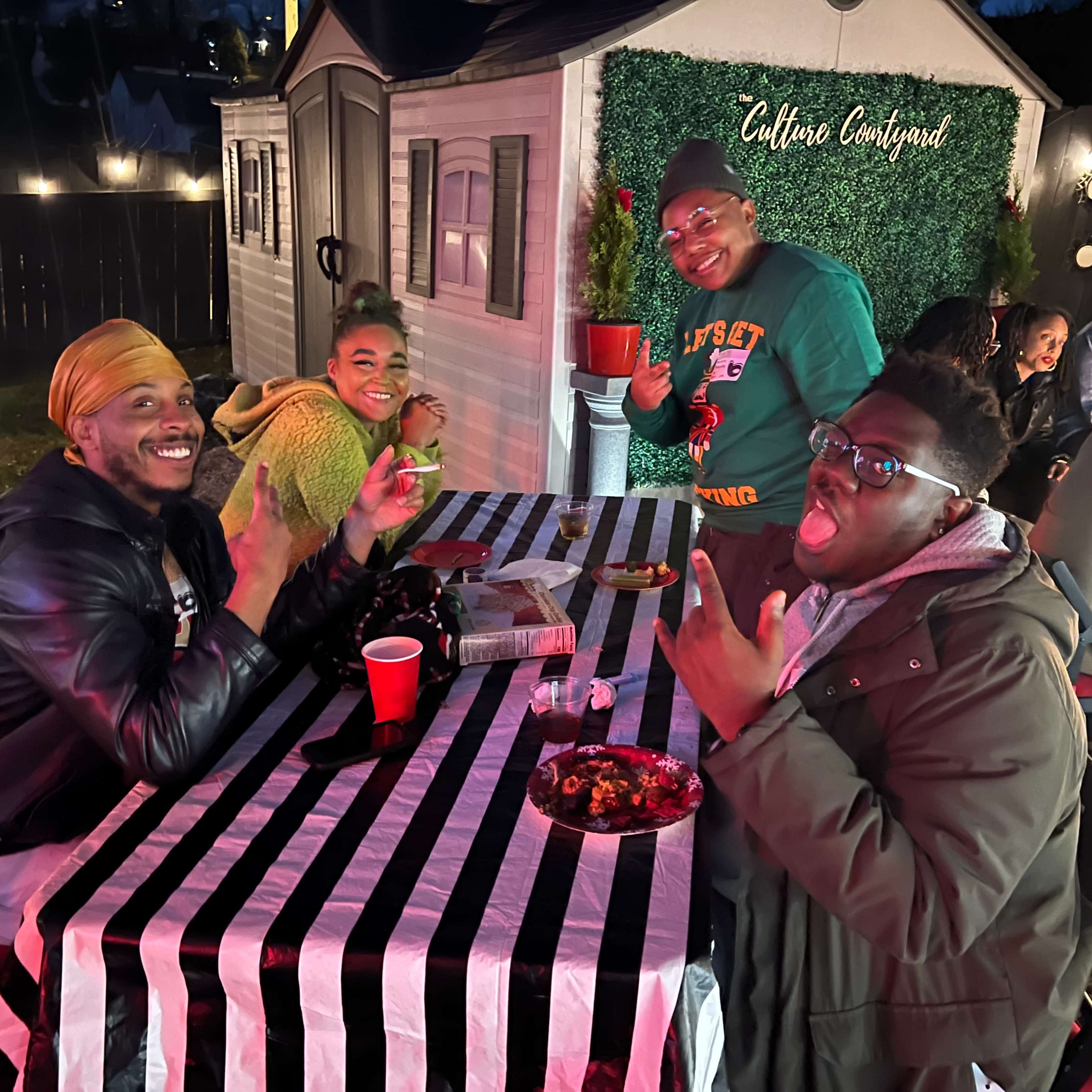 A group of four people gathers around a striped table, smiling and enjoying food at a lively outdoor event.