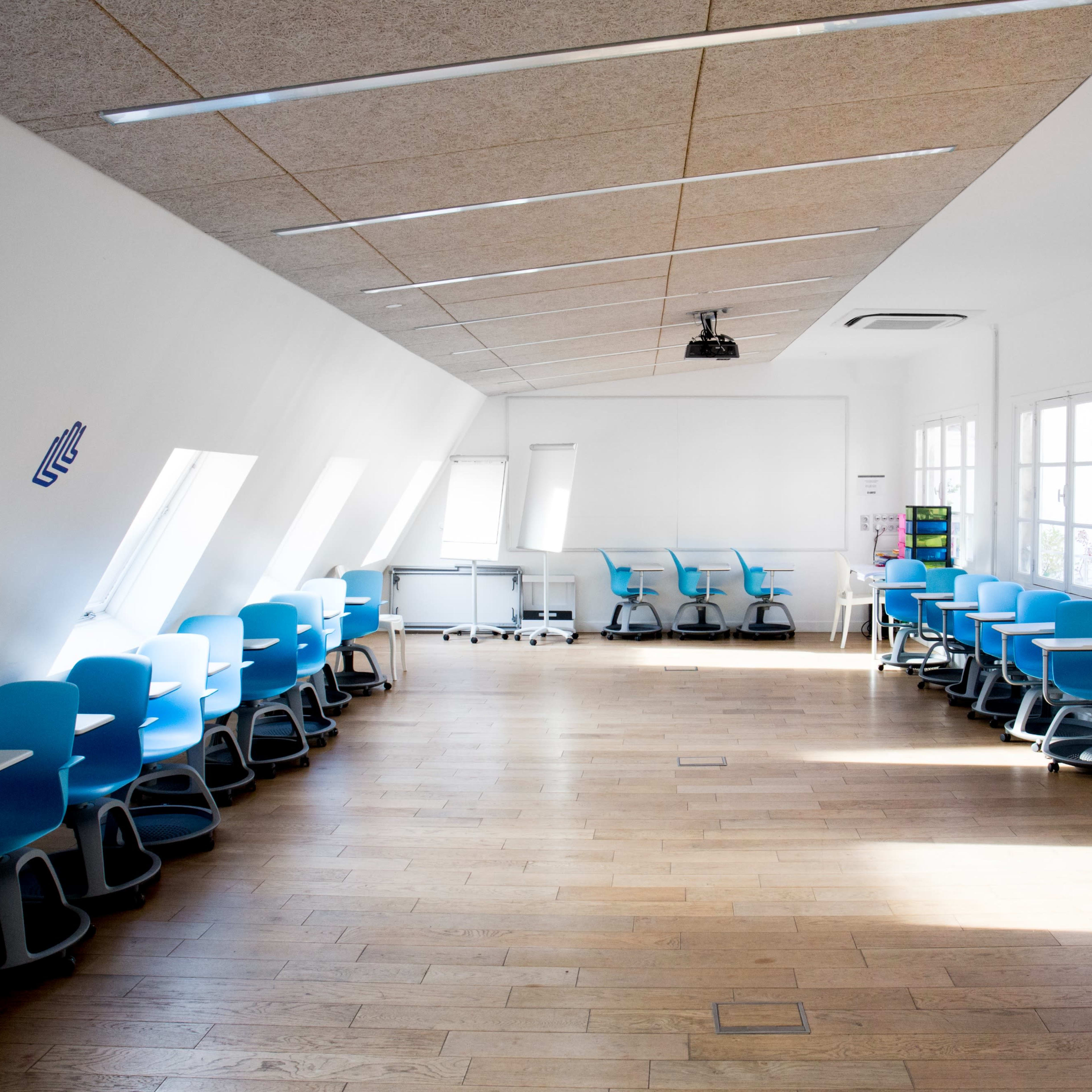 A classroom with blue chairs arranged in rows along the walls and wooden flooring, illuminated by natural light from windows.