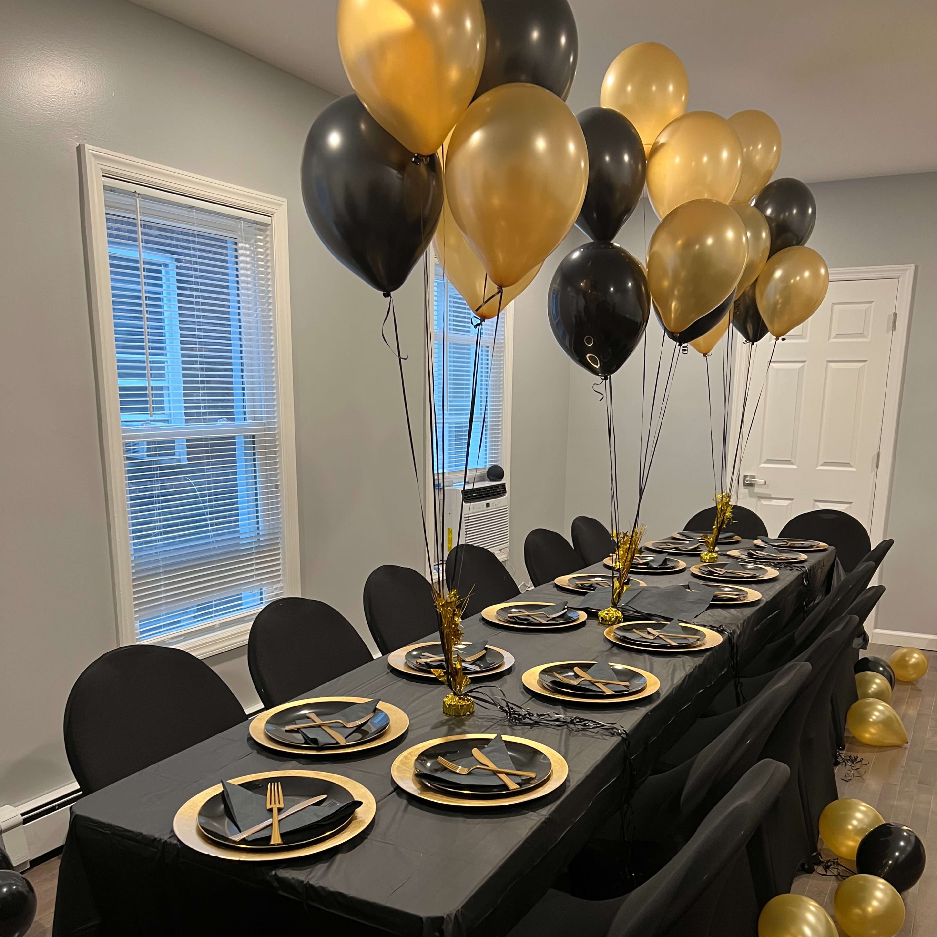 A long dining table is set with black and gold tableware, surrounded by black chairs and adorned with black and gold balloons.