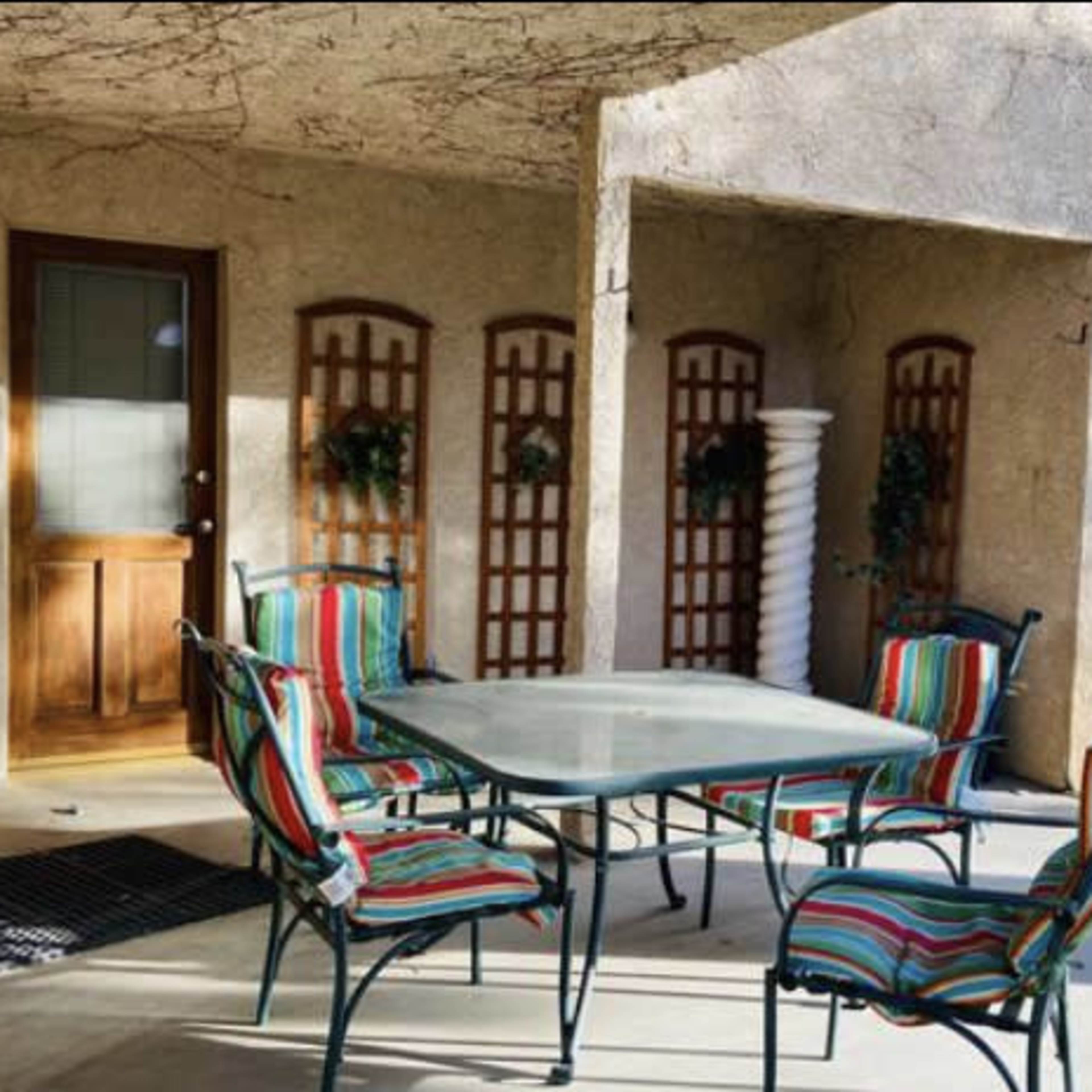 A covered outdoor patio with a glass table and striped chairs, a barbecue grill, and decorative trellises on the walls.