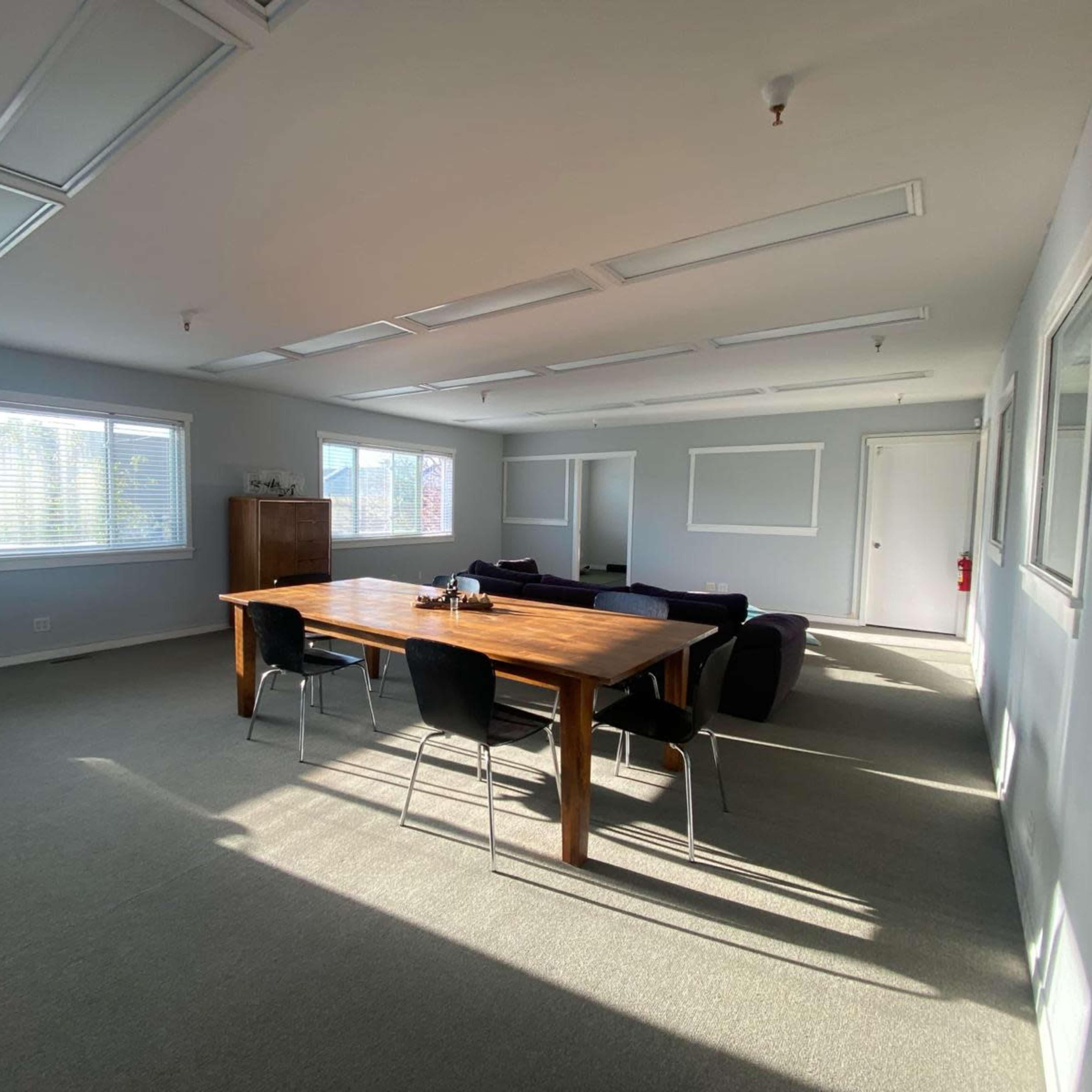 A spacious office features a wooden table surrounded by chairs, with large windows illuminating the carpeted floor.