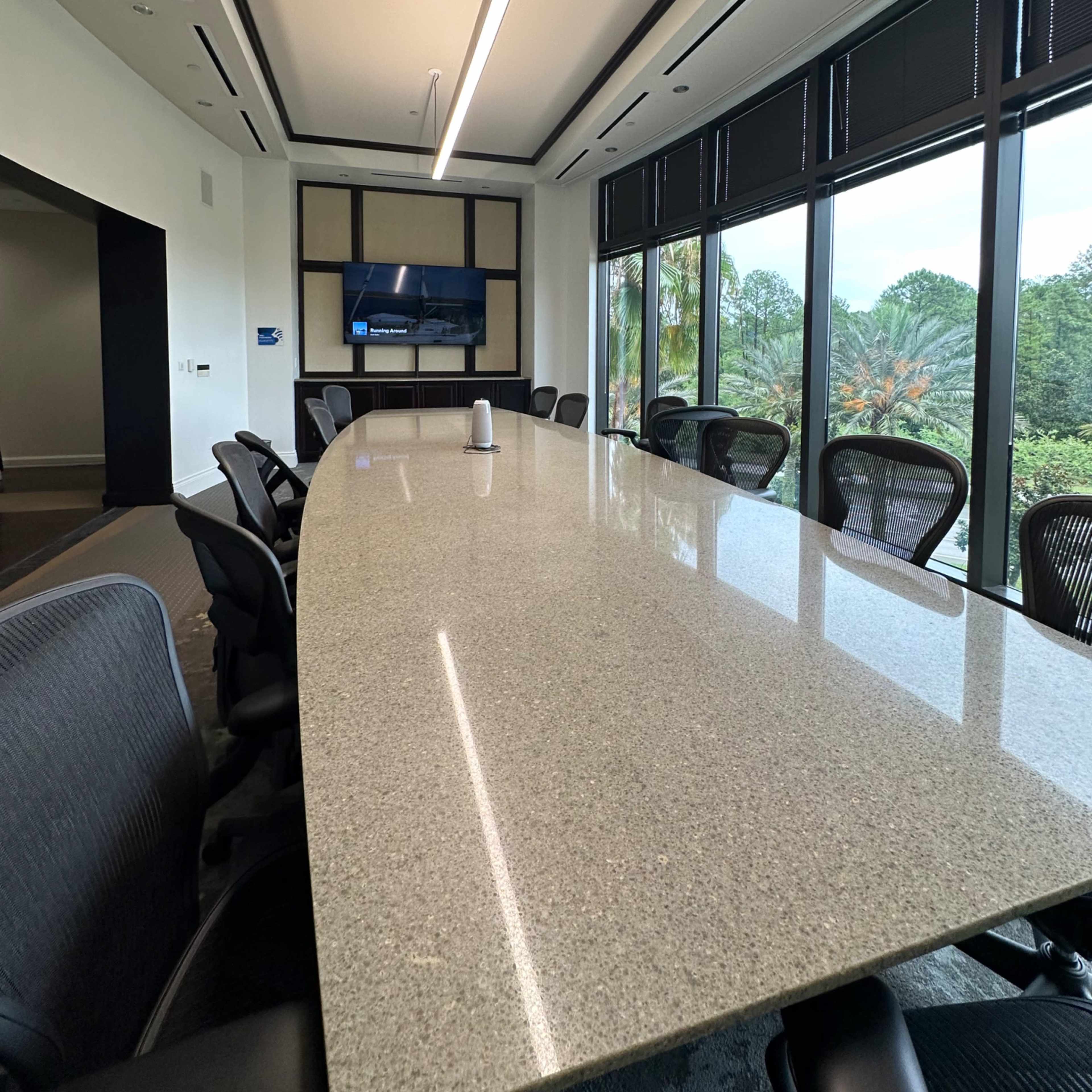 A modern conference room features a long granite table, ergonomic chairs, and large windows overlooking a green landscape.