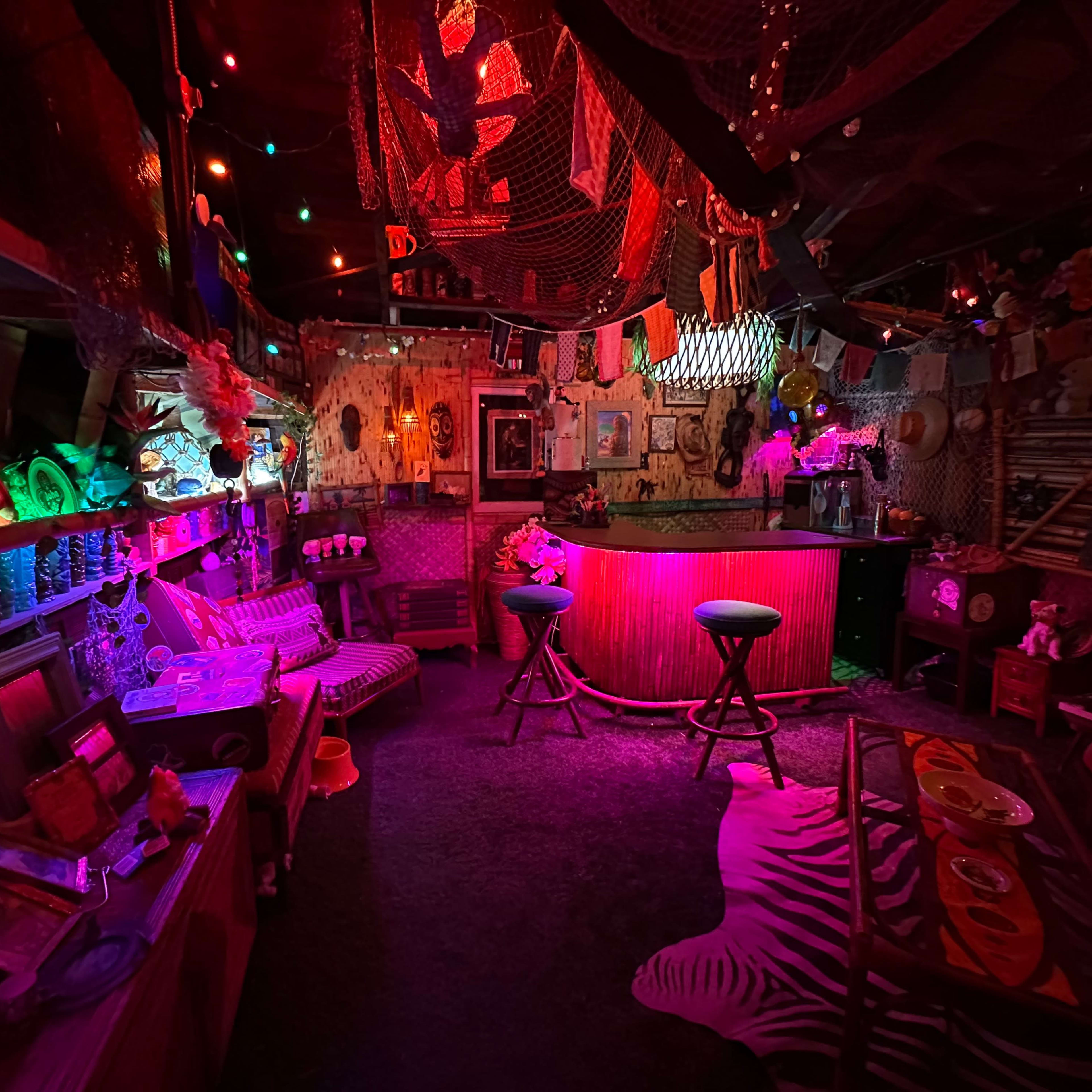 A dimly lit, eclectic bar adorned with colorful lights, decorative nets, and various framed pictures, creating a vibrant and inviting atmosphere.