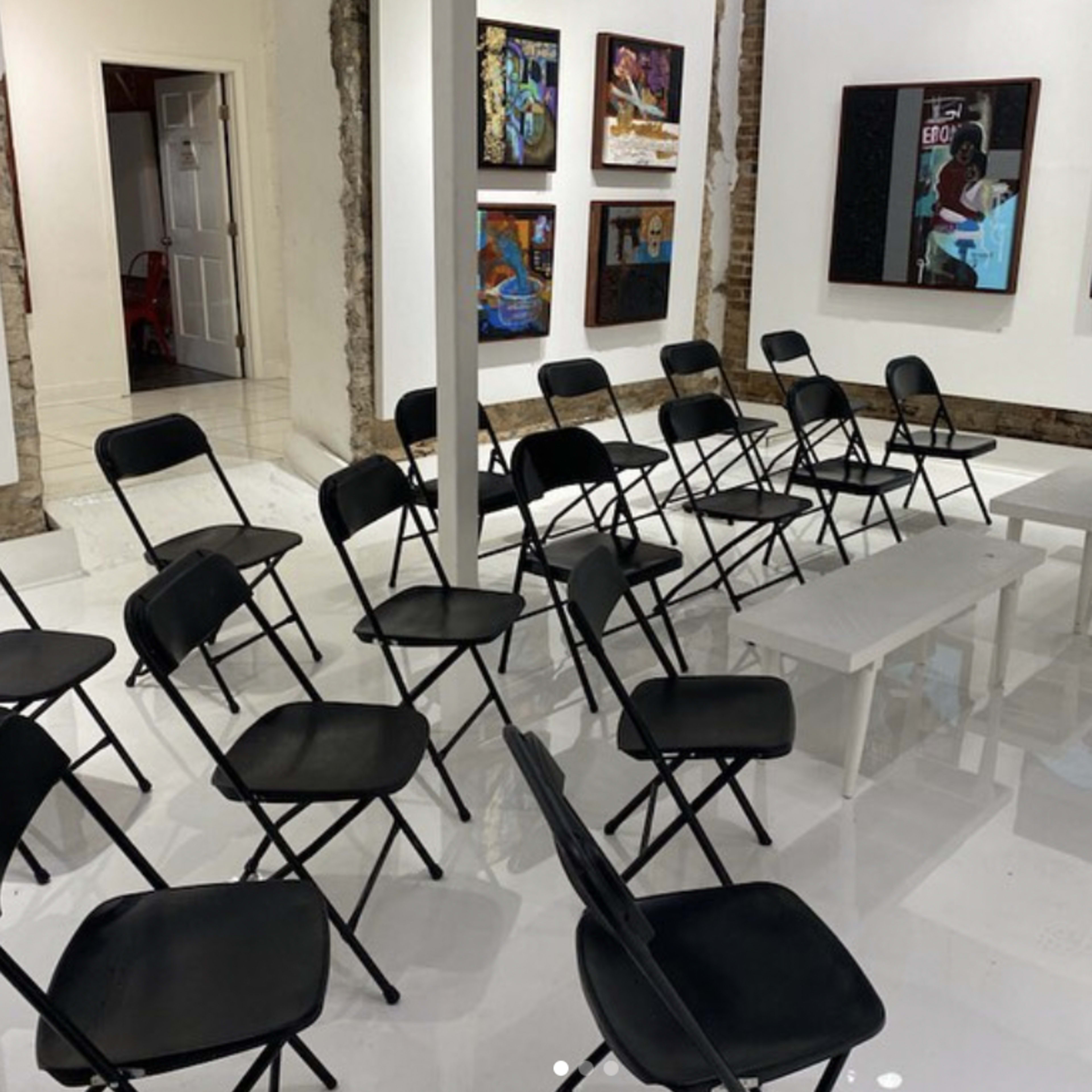A room with several black folding chairs arranged in a circular pattern around a small white table, with artwork displayed on the walls.
