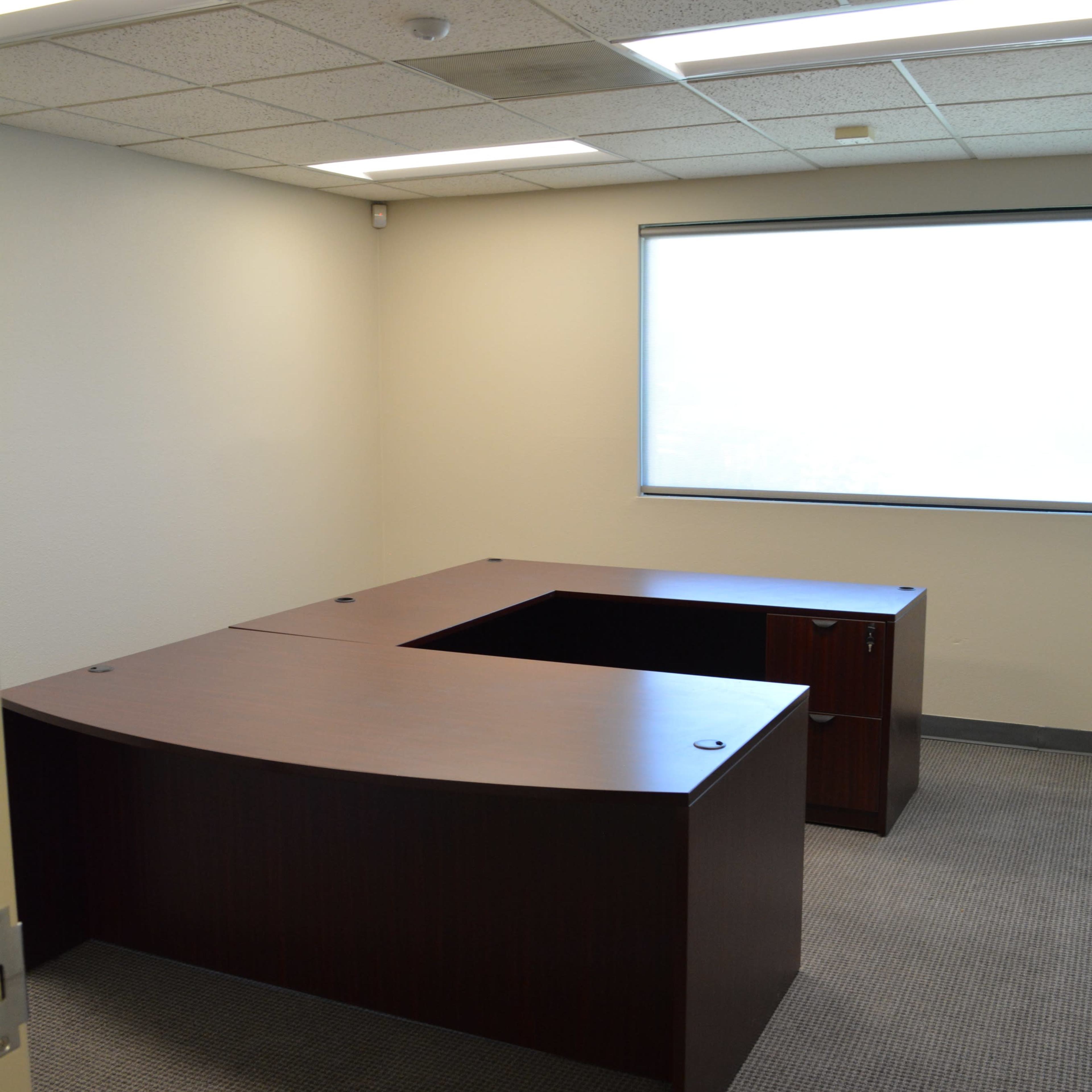 An empty office space featuring two large dark wooden desks arranged in an L-shape, with beige walls and a window allowing natural light.