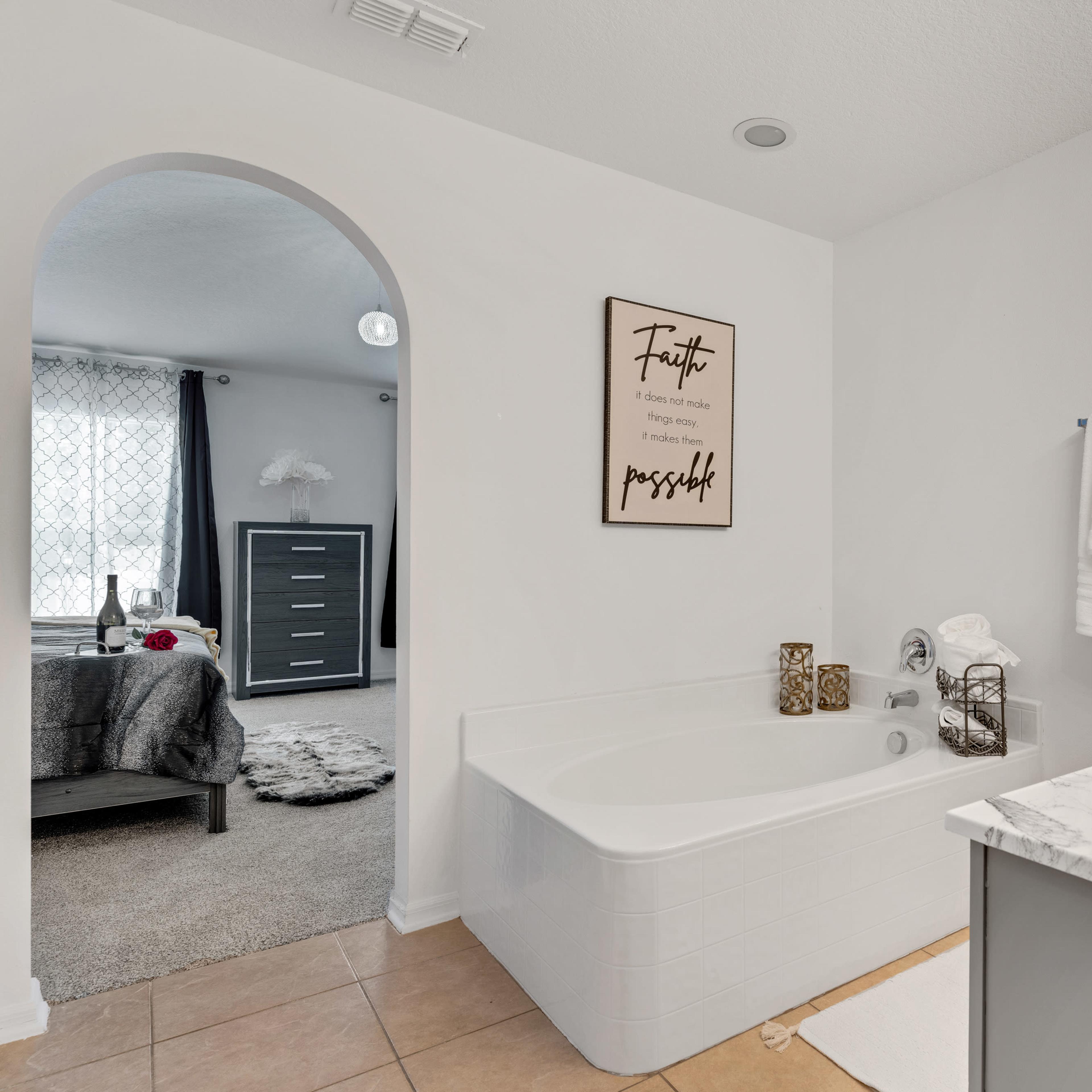 A modern bathroom features a white soaking tub and a marble countertop sink, with a minimalistic decorative poster on the wall and towels neatly arranged nearby, visible through an archway leading to a softly lit bedroom.